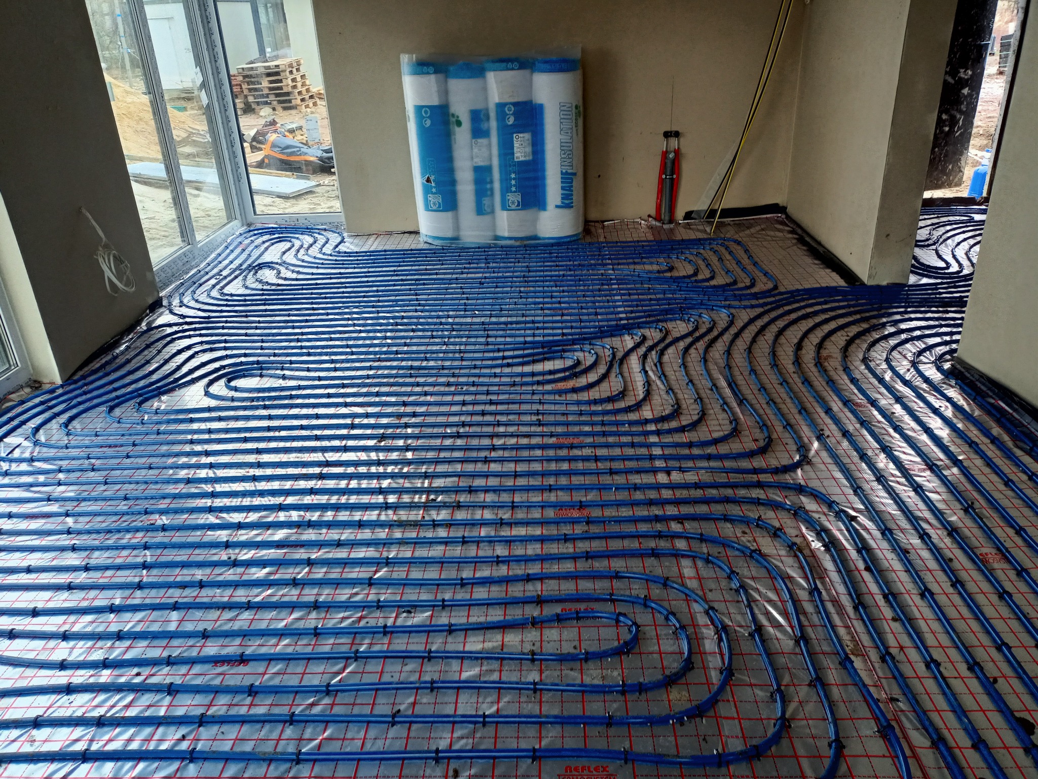 Rozłożone niebieskie rurki na folii z czerwoną siatką, przygotowane do zalania wylewką, w tle rolki izolacji Knauf Insulation i okno z widokiem na plac budowy.