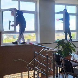 Patriot - Dwóch mężczyzn czyści okna w biurowcu, stojąc na parapecie. Widoczne narzędzia, wiadro i schody. Jasne światło dzienne.