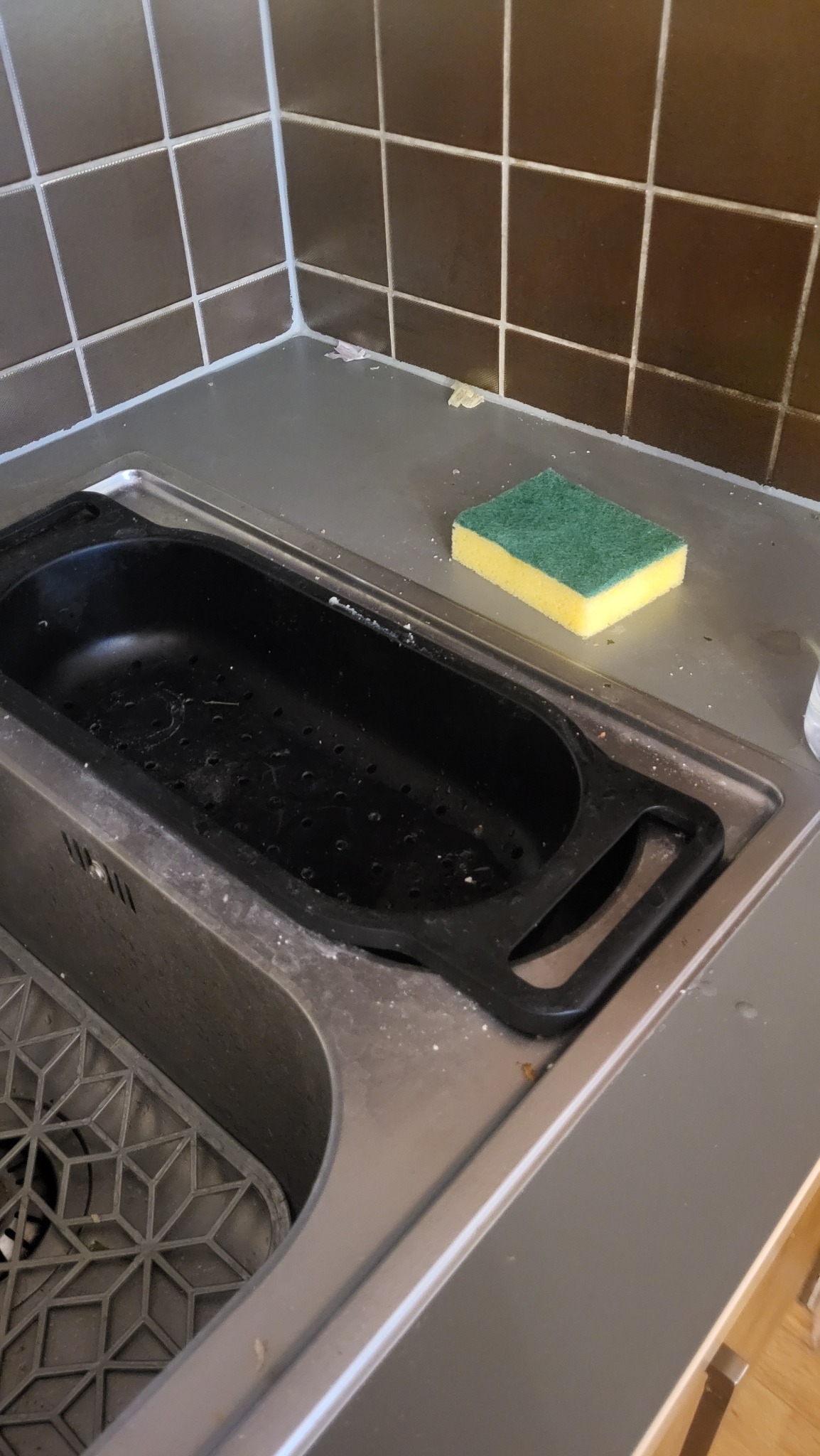 Zlew kuchenny przed czyszczeniem: widoczne zabrudzenia na powierzchni, gąbka leży obok ociekacza. Brązowe płytki w tle.