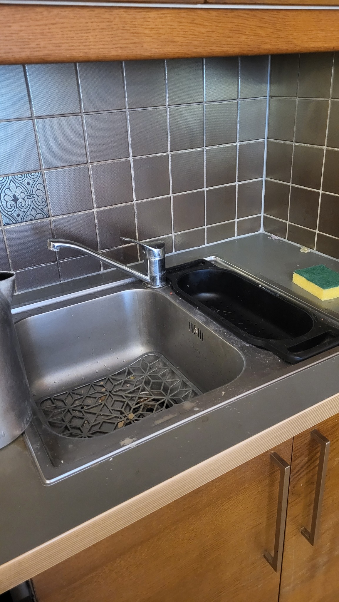 Umywalka kuchenna ze stali nierdzewnej z kranem, czarną wkładką i gąbką. Widoczne brązowe płytki na ścianie i drewniane szafki. Ujęcie z bliska, skupione na detalu.