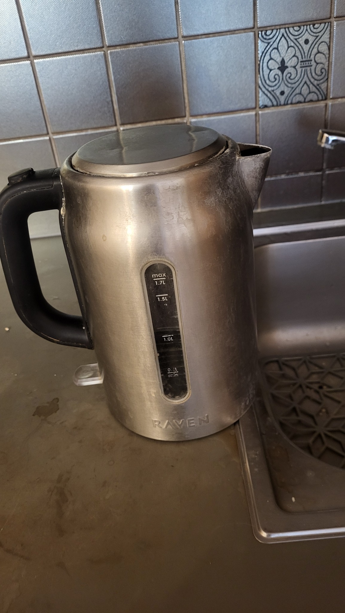 Brudny, metalowy czajnik elektryczny marki Raven stoi na blacie kuchennym obok zlewu. Widoczne zacieki i osad na powierzchni czajnika. Tło stanowią szare płytki.