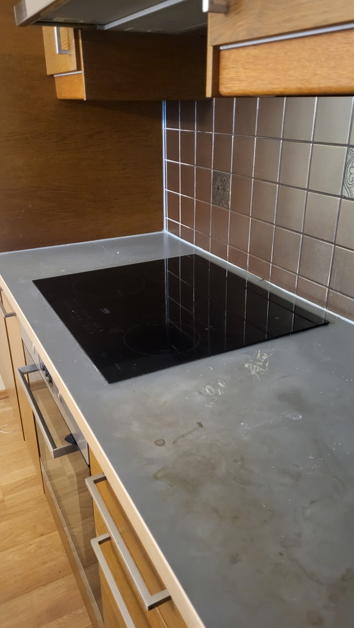 Zabrudzony blat kuchenny z płytą indukcyjną, widoczne plamy i zacieki. Tło stanowią brązowe płytki i drewniane szafki. Ujęcie z bliska, ukazujące stopień zabrudzenia.