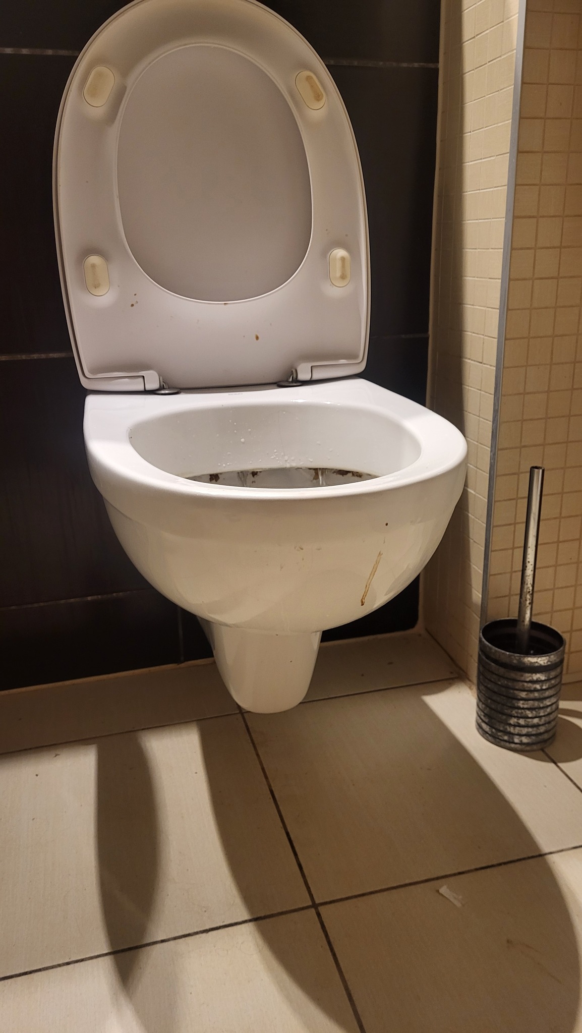 Z bliska: brudna toaleta z otwartą klapą i szczotką. Widoczne zabrudzenia na ceramice i podłodze. Niefortunny kąt i oświetlenie.