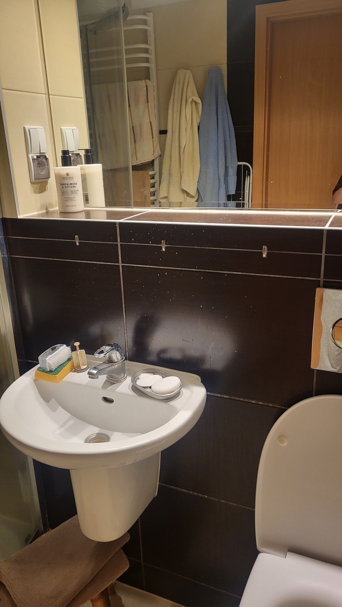 Umywalka z baterią i mydłem w łazience z ciemnymi płytkami, widoczne ślady po zaschniętej wodzie. W tle lustro z odbiciem ręczników i grzejnika.