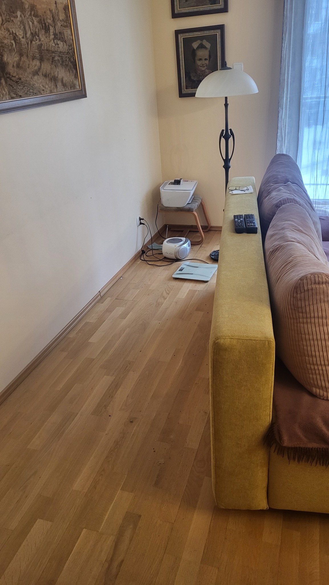 Parkiet z drobnymi zabrudzeniami w rogu pokoju, obok wagi łazienkowej i odkurzacza. W tle sofa, lampa i obrazy na ścianie. Widok z góry.