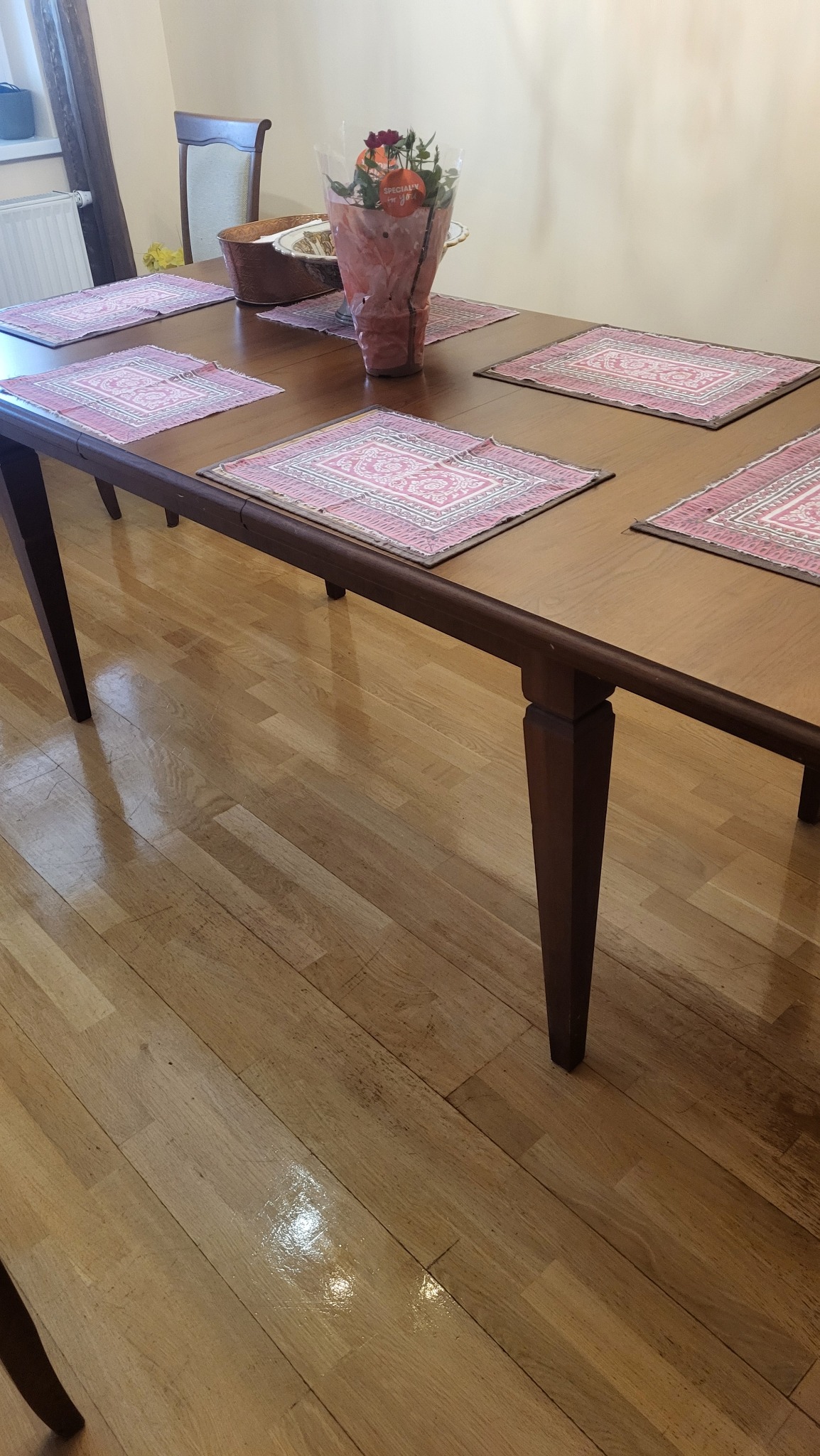 Wypolerowany stół z dekoracyjnymi podkładkami i bukietem kwiatów, na tle lśniącej drewnianej podłogi. Jasne wnętrze po sprzątaniu.