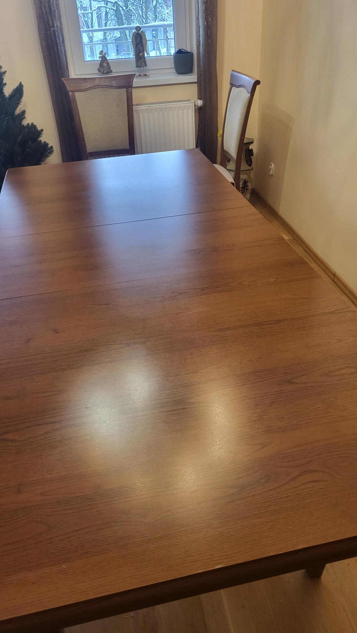 Wypolerowany, drewniany stół w jadalni po czyszczeniu, z widocznymi refleksami światła. W tle okno z dekoracjami i krzesła.
