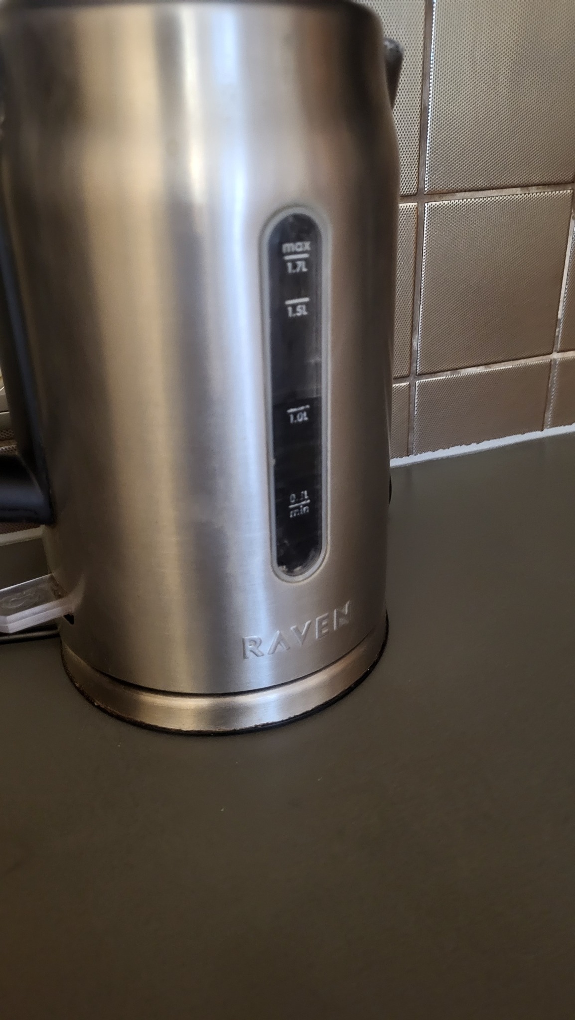 Błyszczący, metalowy czajnik elektryczny marki Raven na blacie kuchennym, z widocznym wskaźnikiem poziomu wody i tłem z płytek. Minimalistyczny design.