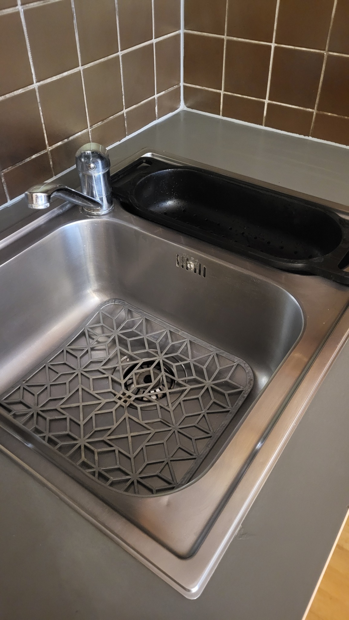Błyszczący, stalowy zlew kuchenny z szarą, geometryczną wkładką i chromowaną baterią na tle brązowych płytek. Widoczna czarna ociekarka.