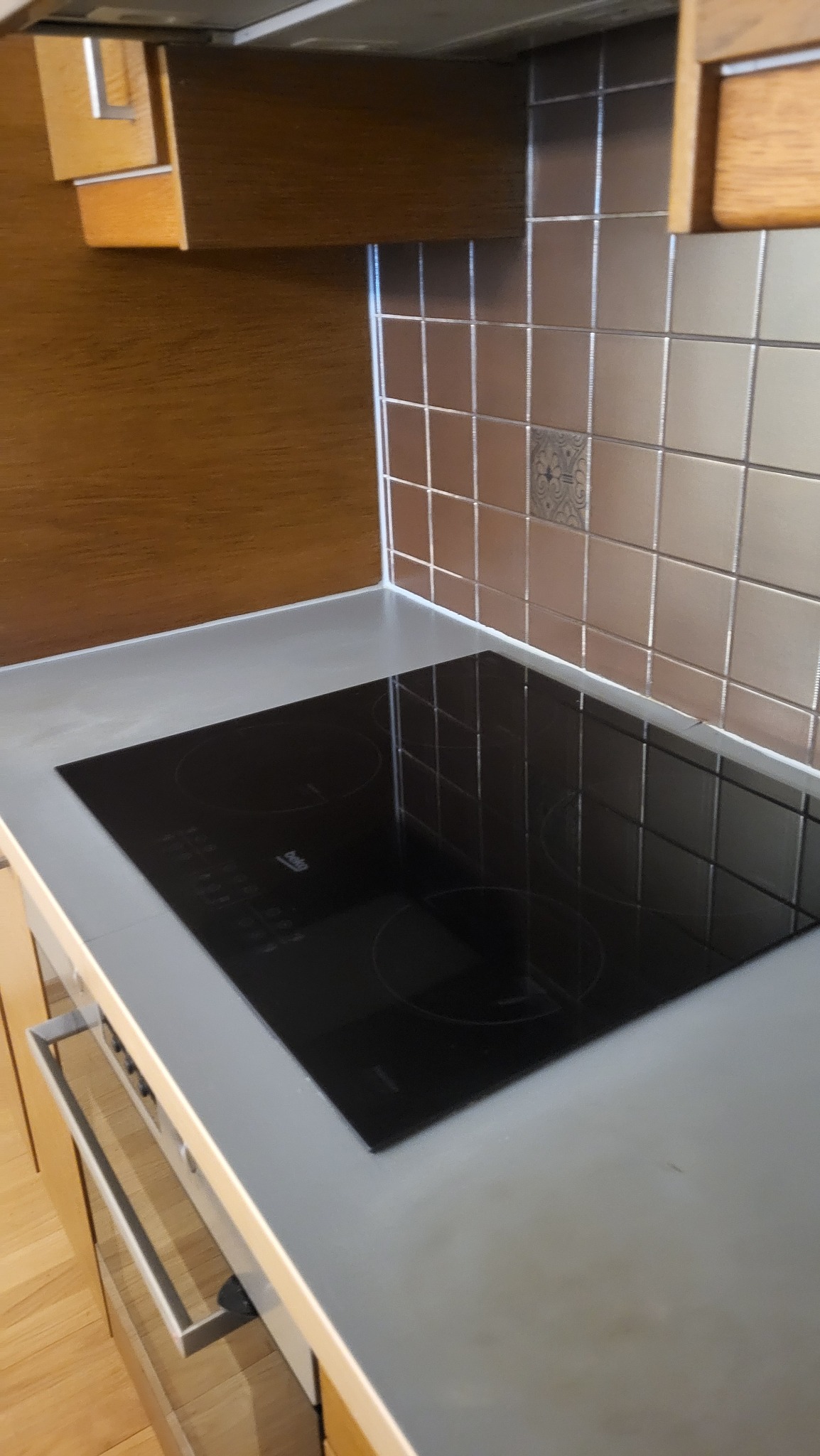 Błyszcząca, czarna płyta indukcyjna marki Beko, osadzona w szarym blacie kuchennym, z tłem z płytek w odcieniach szarości i brązu. Widok z góry, ukośnie.