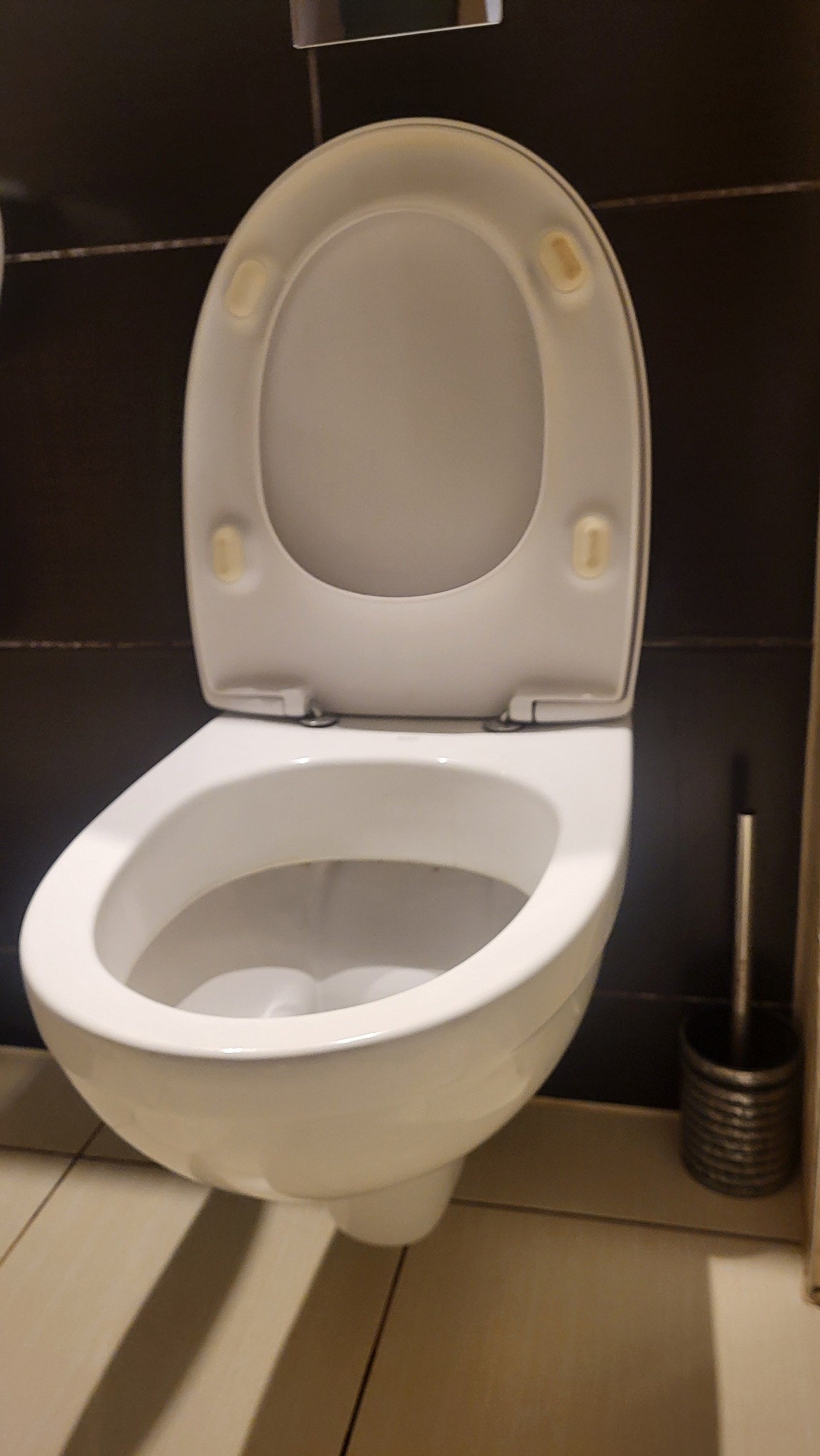 Biała, otwarta muszla klozetowa z deską na tle ciemnych płytek i beżowej podłogi. Widoczna szczotka do WC. Precyzyjne czyszczenie toalety w łazience.