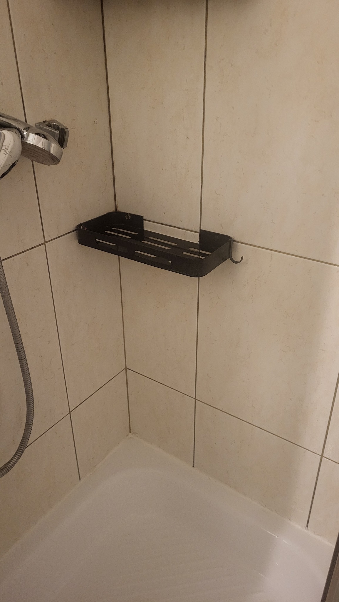 Półka łazienkowa narożna, czarna, zamontowana na jasnych, kwadratowych płytkach w kabinie prysznicowej. Widoczny fragment słuchawki prysznicowej i brodzika.