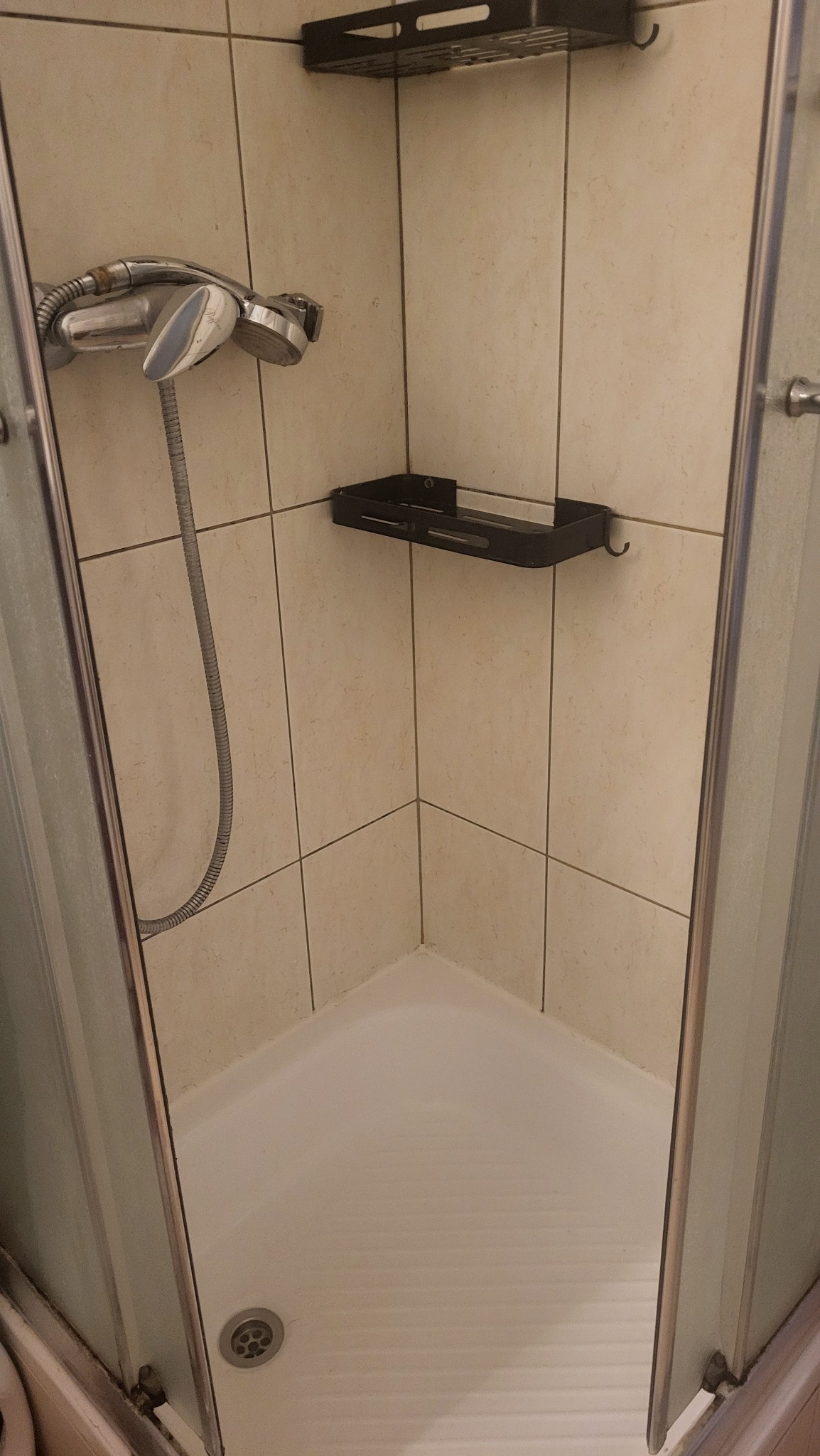 Wnętrze kabiny prysznicowej z jasnymi płytkami, metalową słuchawką i półkami. Widoczny odpływ w brodziku. Zdjęcie z perspektywy wnętrza kabiny.