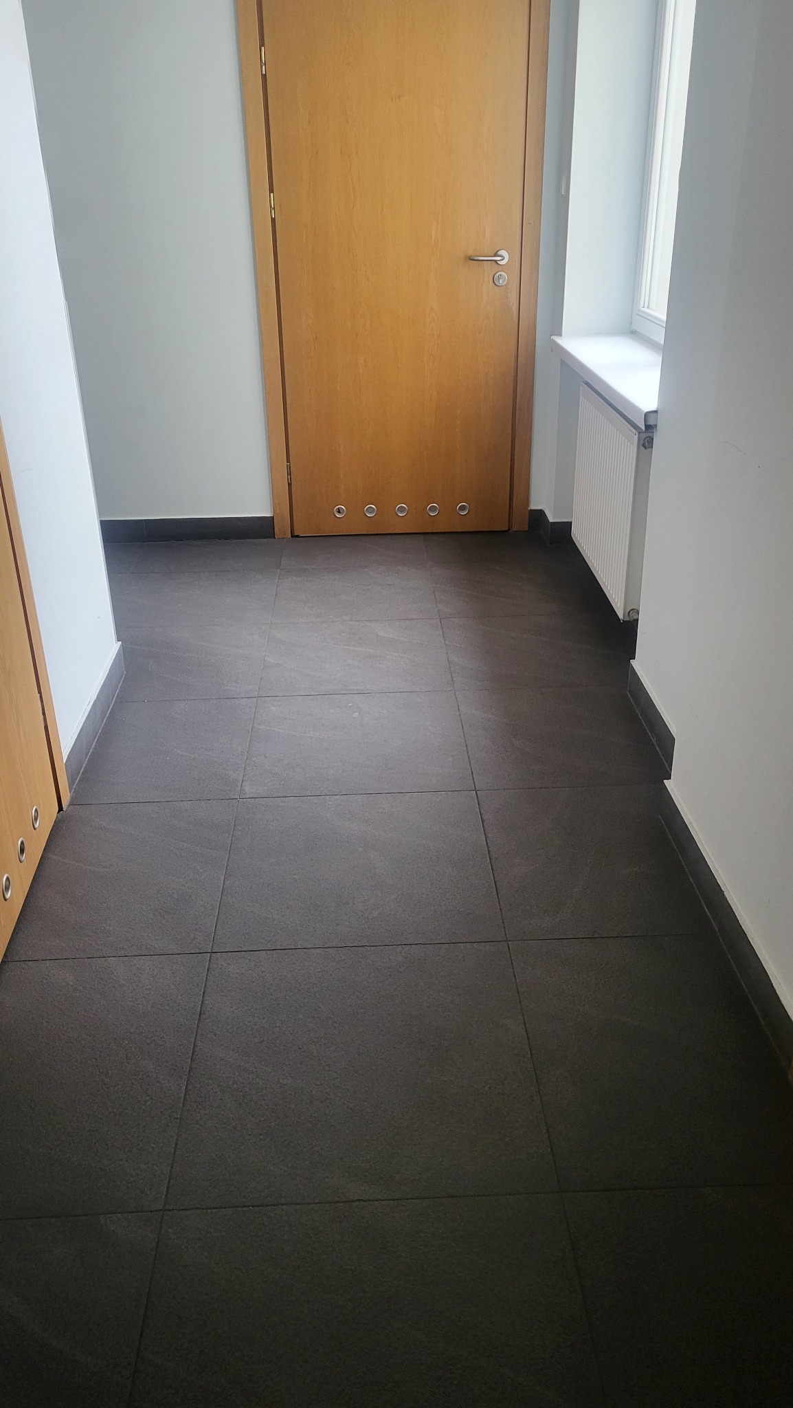 Ciemne, kwadratowe płytki podłogowe w korytarzu, widok z góry. W tle drewniane drzwi z metalowymi detalami i biały grzejnik pod oknem. Minimalistyczny design.
