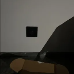 Kamera alarmowa w czarnej obudowie zamontowana na białej ścianie, z widocznym kartonowym pudełkiem na pierwszym planie i fragmentem czarnego przedmiotu po prawej stronie.