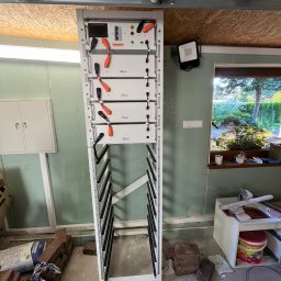 ELEKTRO-CIERBIKOWSKI KRYSTIAN CIERBIKOWSKI - Biała szafa z modułami akumulatorów Deye w garażu, widoczne kable z pomarańczowymi końcówkami, na podłodze worki z zaprawą i cegły, w tle okno z widokiem na ogród.