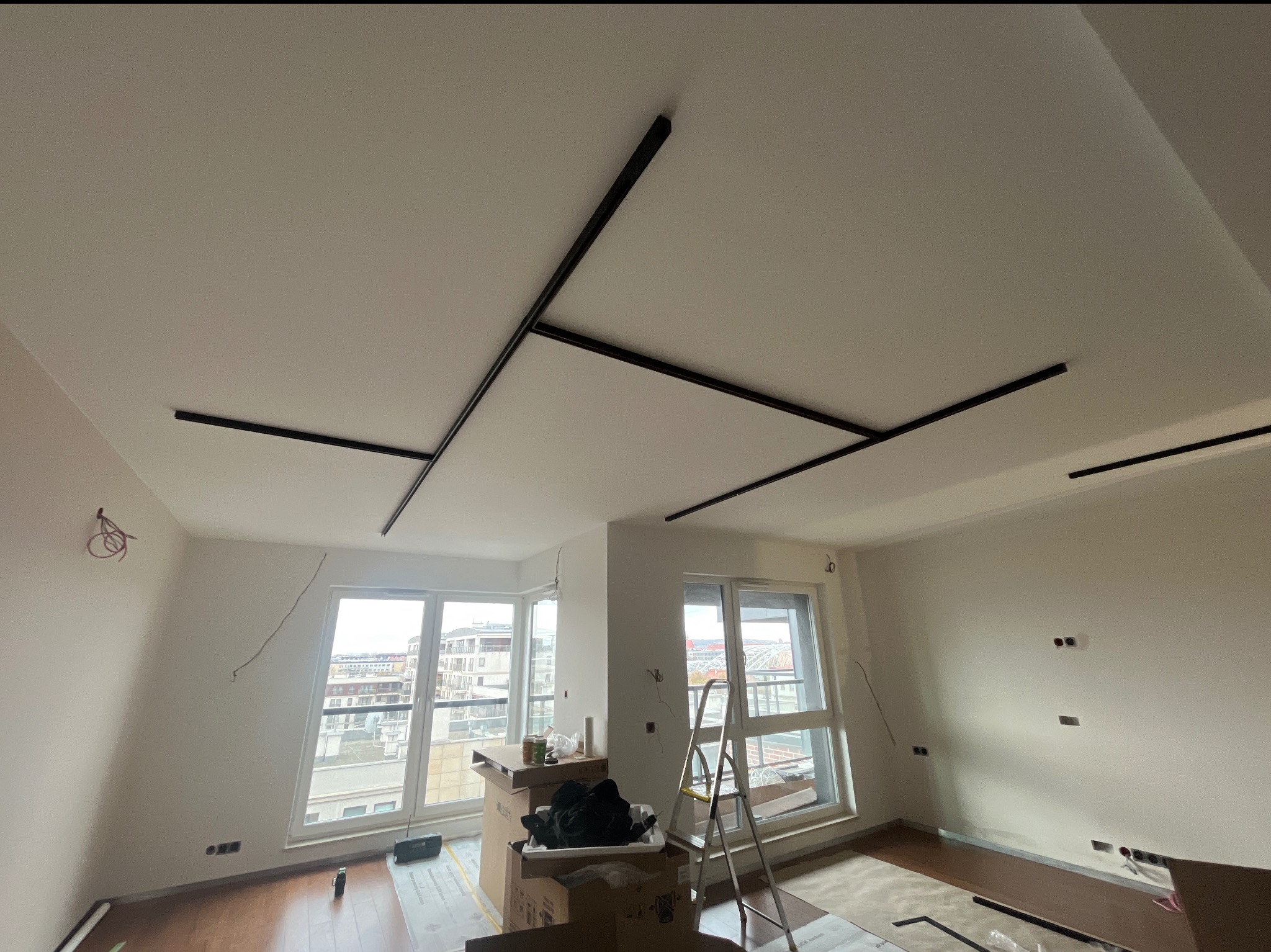 Nowoczesny system oświetlenia sufitowego z czarnymi, prostymi oprawami w jasnym wnętrzu w trakcie montażu, widoczne przewody elektryczne na ścianach i drabina.