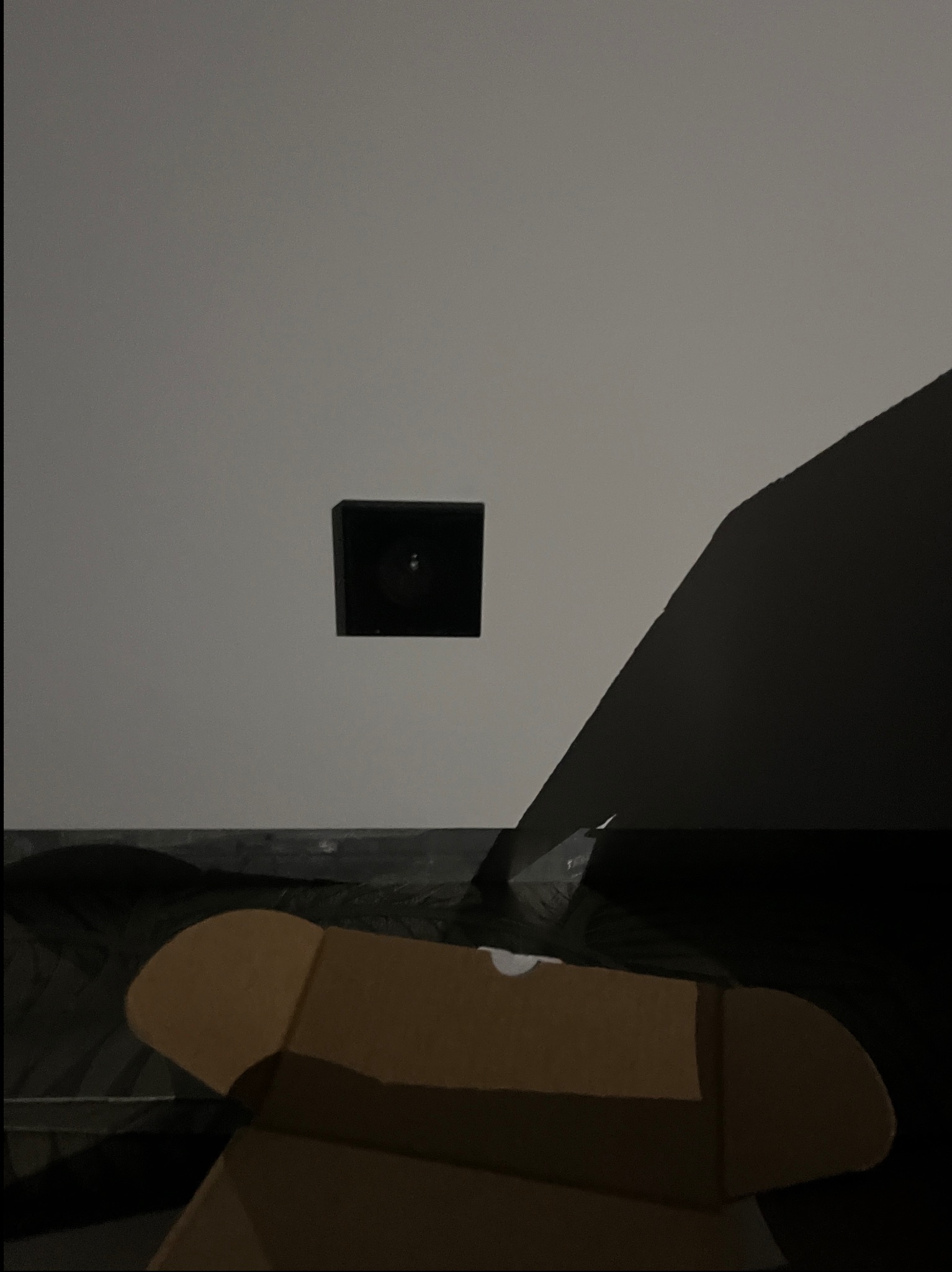 Kamera alarmowa w czarnej obudowie zamontowana na białej ścianie, z widocznym kartonowym pudełkiem na pierwszym planie i fragmentem czarnego przedmiotu po prawej stronie.