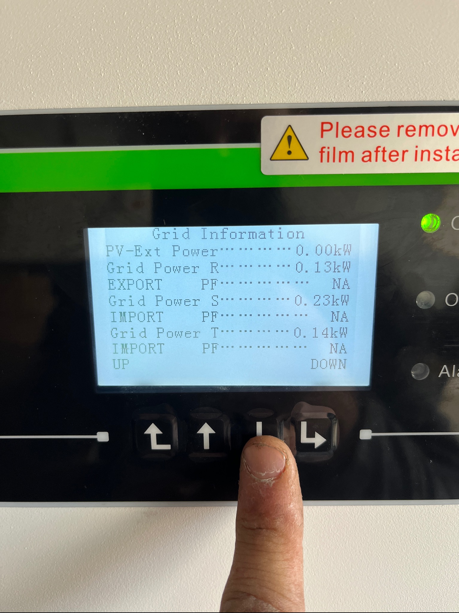 Palec wskazujący na ekranie falownika fotowoltaicznego wyświetlającym dane o produkcji energii i imporcie/eksporcie do sieci, z naklejką ostrzegawczą o usunięciu folii po instalacji.