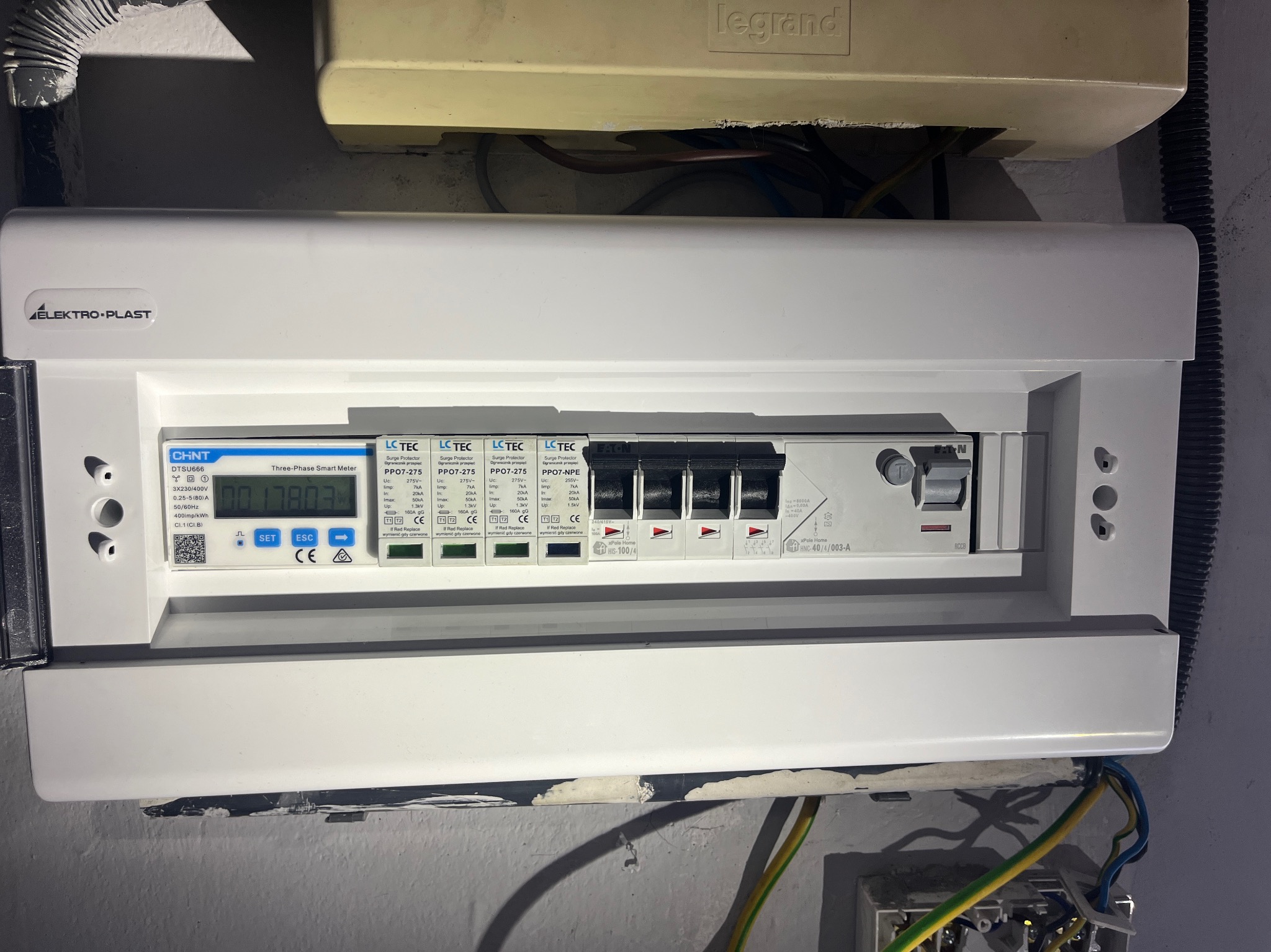 Rozdzielnica elektryczna z licznikiem trójfazowym i zabezpieczeniami przepięciowymi marek Elektro-Plast, CHNT, LC TEC i Eaton, zamontowana na ścianie z widocznymi przewodami elektrycznymi.
