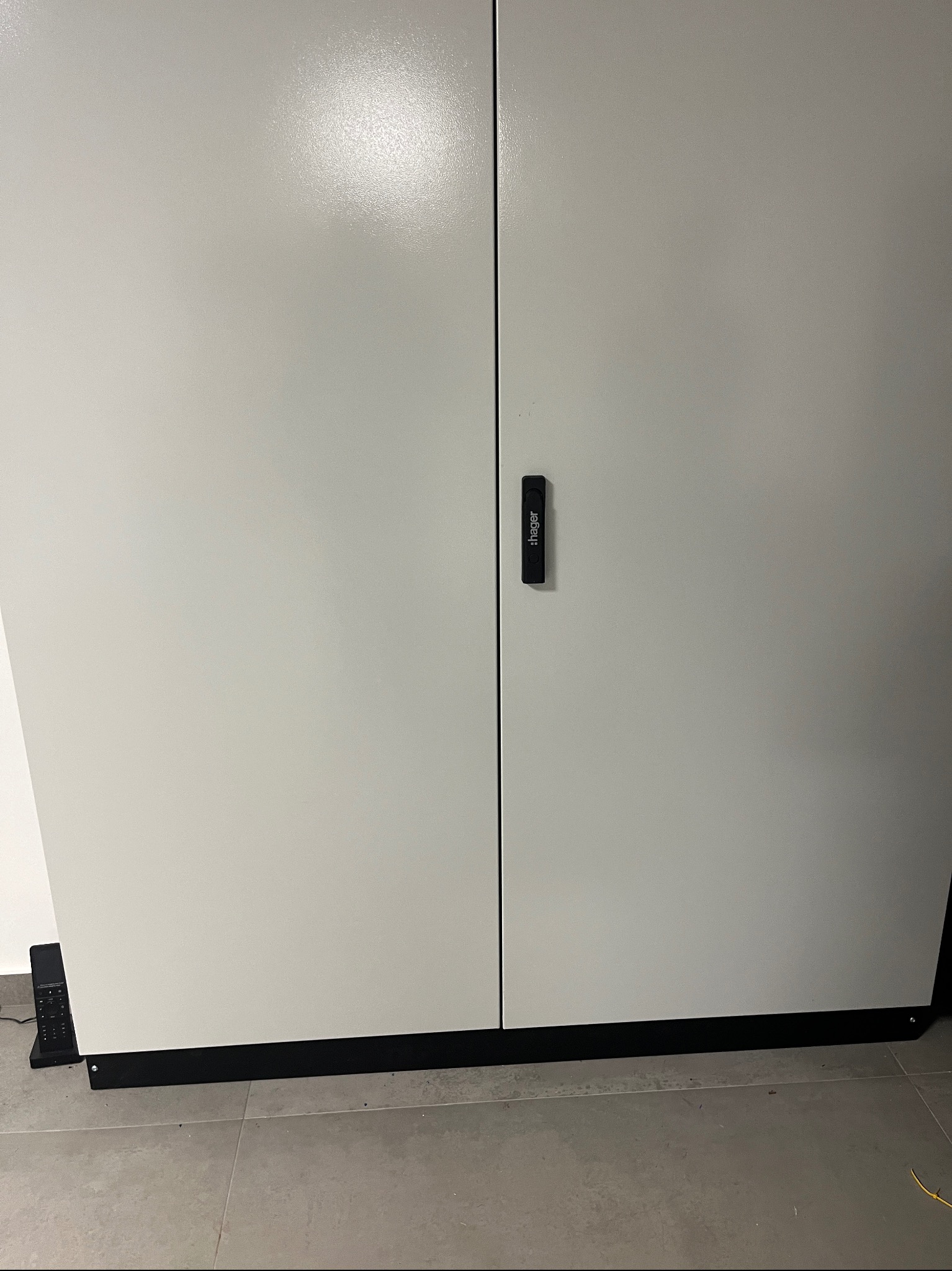 Zamknięta, szara szafa elektryczna firmy Hager z czarnym uchwytem, widoczny pilot alarmowy na podłodze