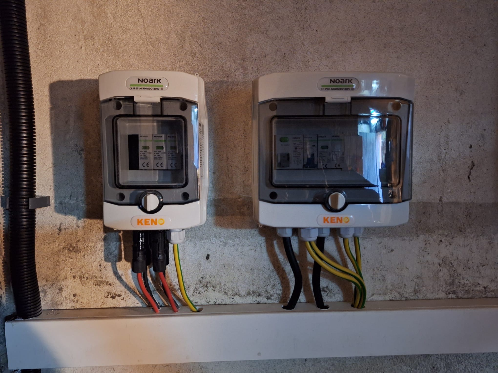 Dwa białe rozdzielacze elektryczne marki Noark, model KENO, zamontowane na szarej, nierównej ścianie z widocznymi przewodami w kolorach czarnym, czerwonym i żółto-zielonym, umieszczone na białej...