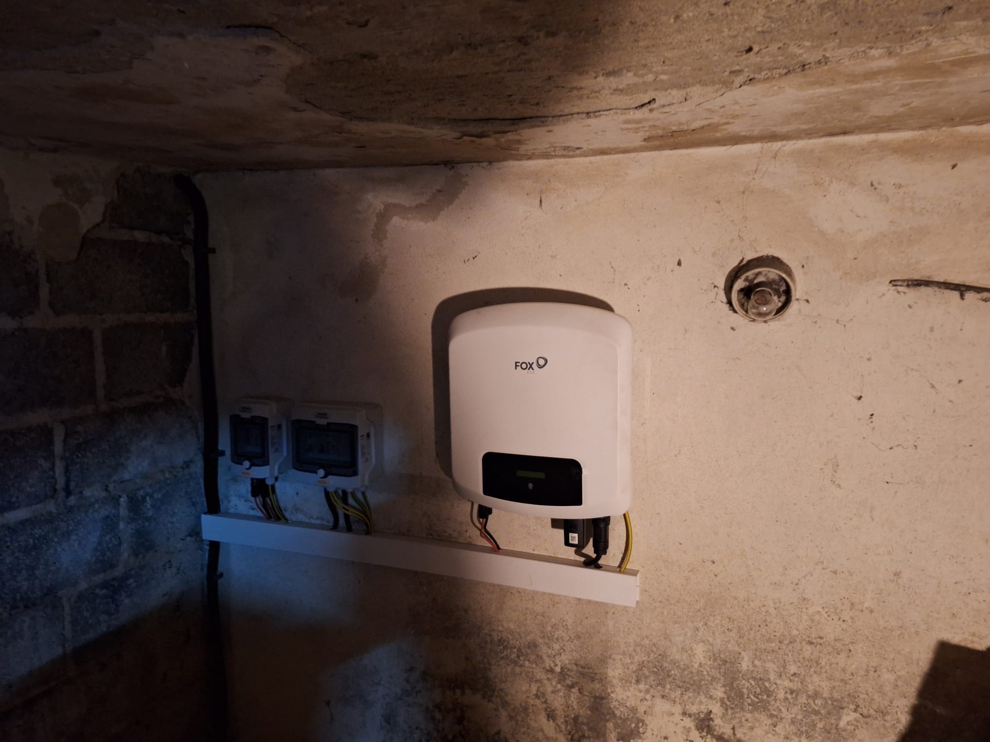 Biały inwerter fotowoltaiczny marki FOX zamontowany na ścianie w piwnicy, obok dwa liczniki elektryczne na białej listwie, widoczne przewody.