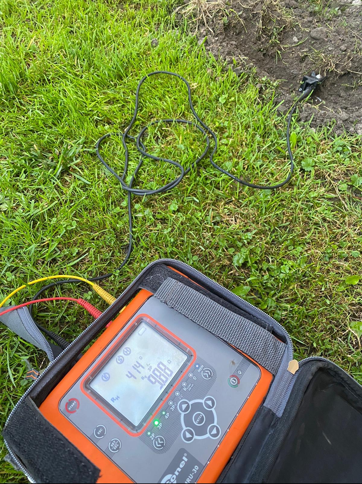Miernik rezystancji uziomu Sonel MRU-30 w otwartym futerale na trawie, z widocznymi przewodami pomiarowymi i sondą wbita w ziemię, podczas pomiaru rezystancji.