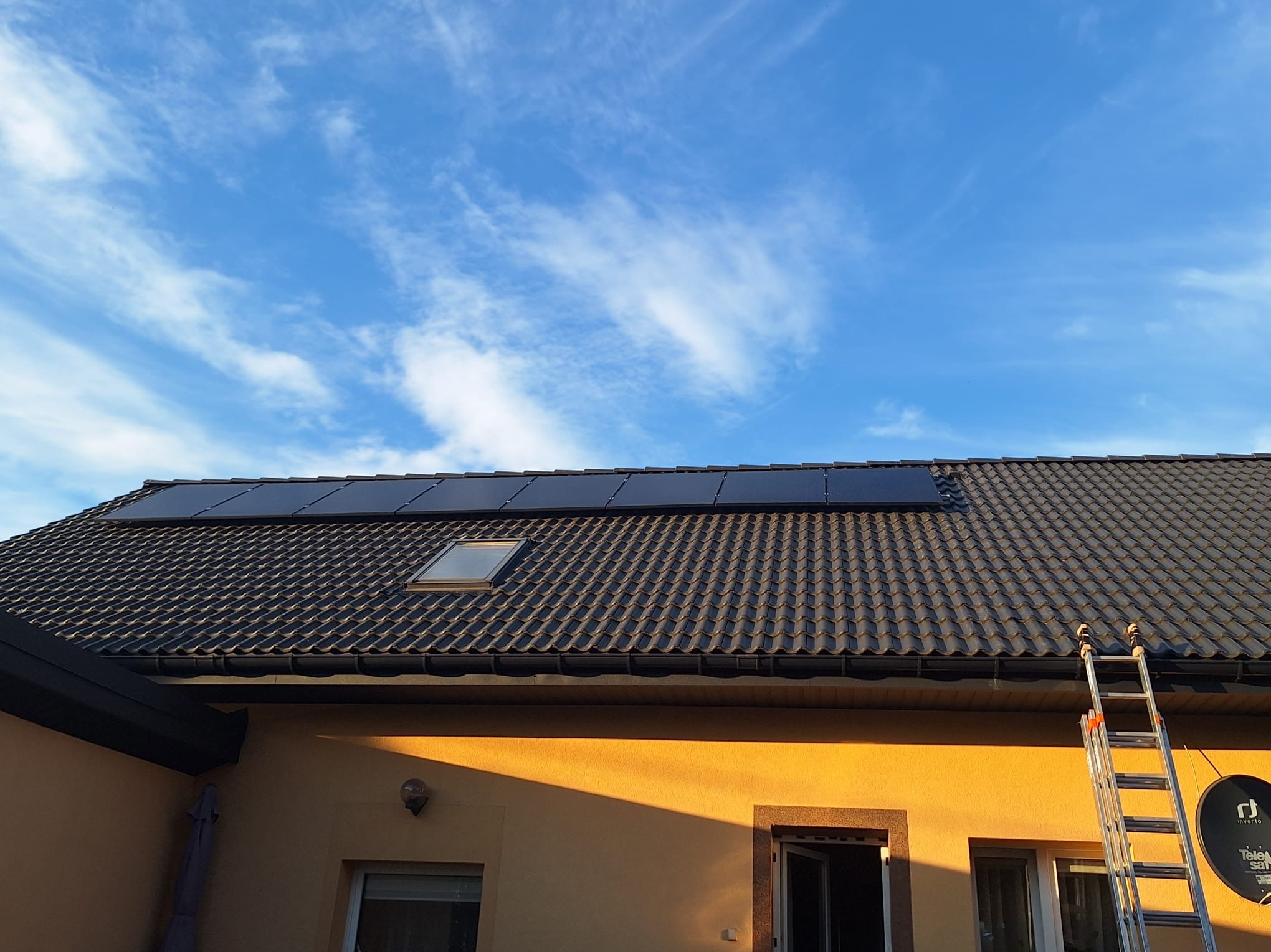 Panele słoneczne na dachu domu z brązową dachówką, widoczne okno dachowe, drabina oparta o ścianę i antena satelitarna, błękitne niebo w tle.