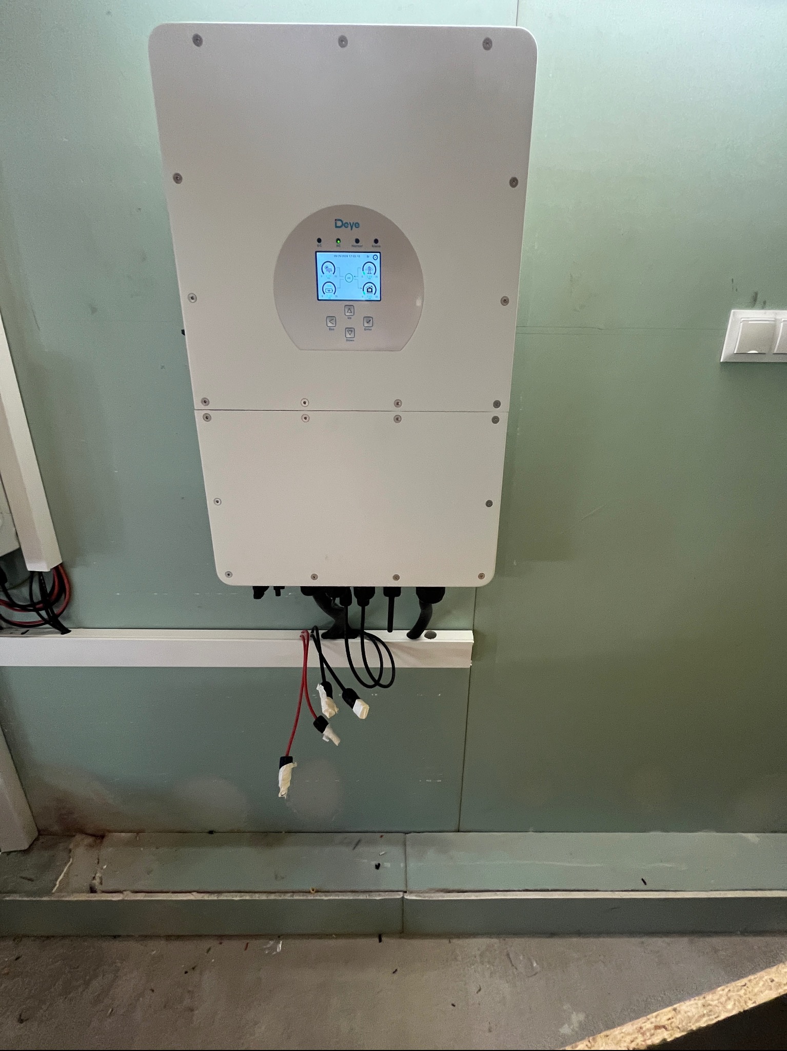 Biały falownik marki Deye zamontowany na zielonej ścianie, widoczne kable z końcówkami MC4, ekran falownika z ikonami i wskaźnikami.