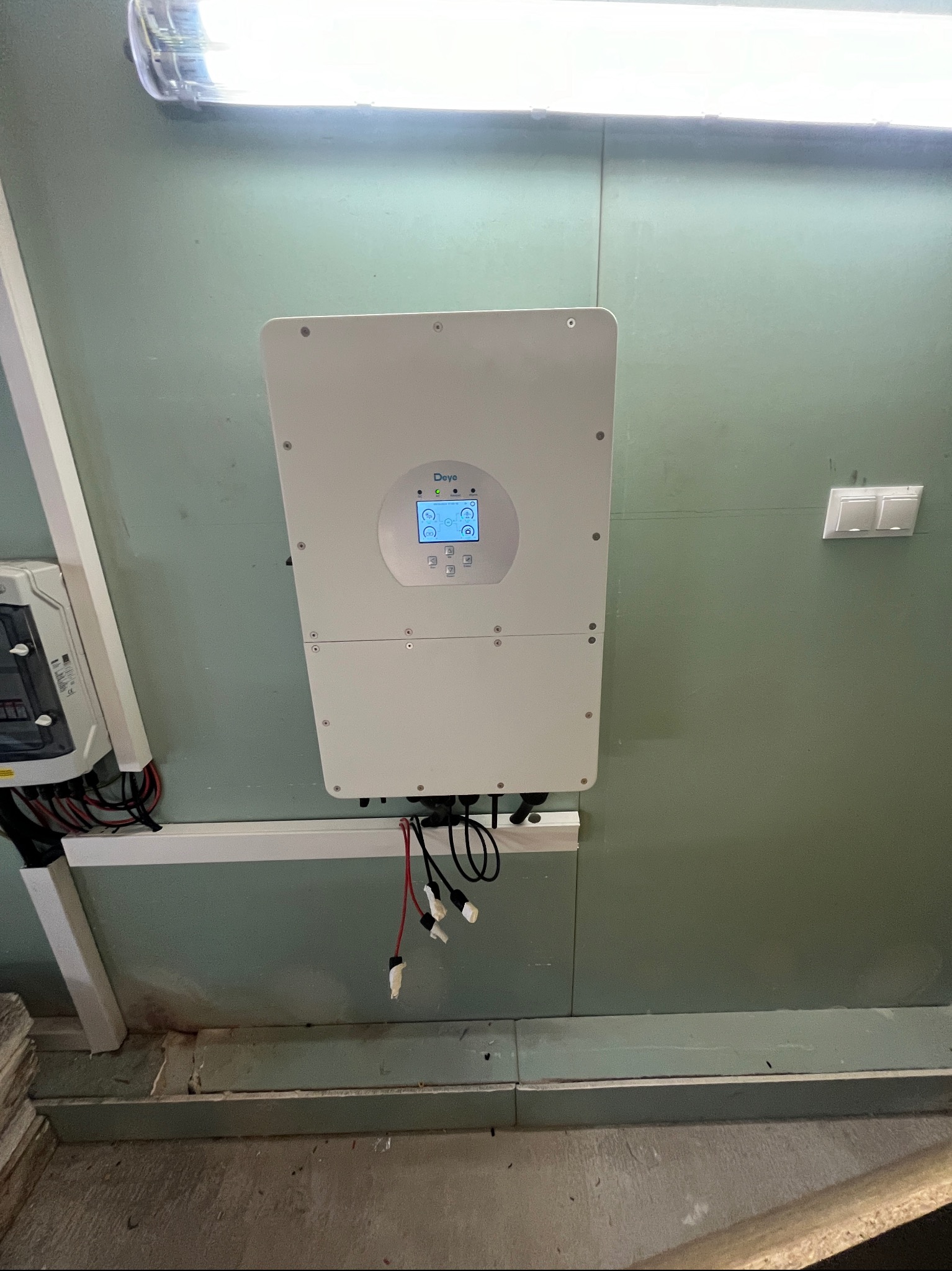 Zamontowany biały falownik Deye z podłączonymi kablami, widoczne także zabezpieczenia elektryczne na ścianie pokrytej zieloną farbą i oświetlenie LED.