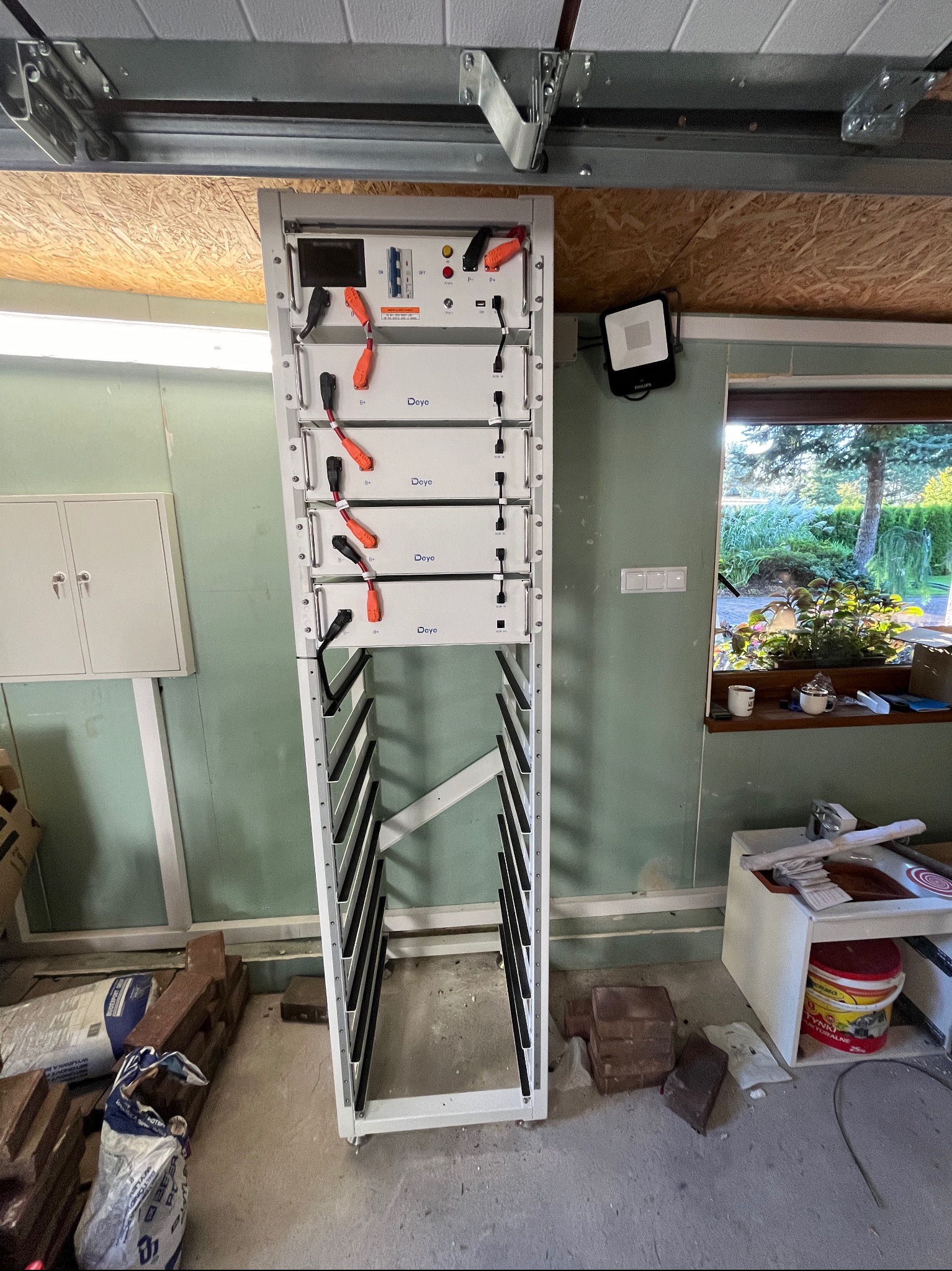 Biała szafa z modułami akumulatorów Deye w garażu, widoczne kable z pomarańczowymi końcówkami, na podłodze worki z zaprawą i cegły, w tle okno z widokiem na ogród.