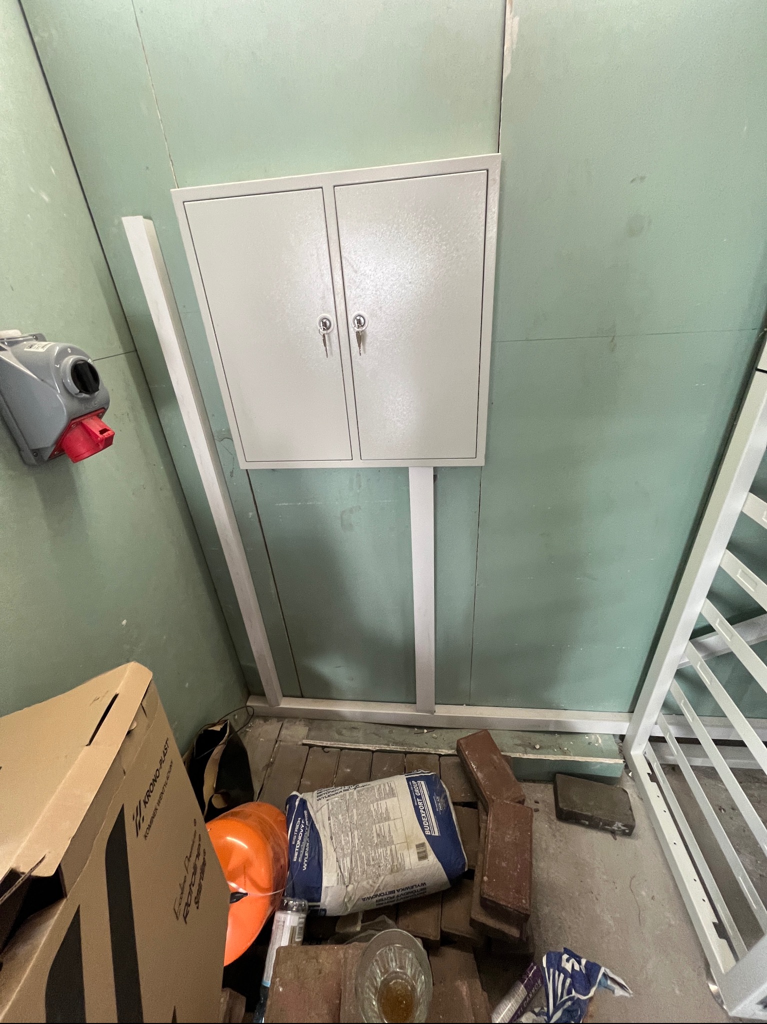 Szara skrzynka elektryczna zamykana na klucz, zamontowana na zielonej ścianie z płyt gipsowo-kartonowych, z widocznymi elementami instalacyjnymi i budowlanymi: puszka elektryczna z czerwonym...