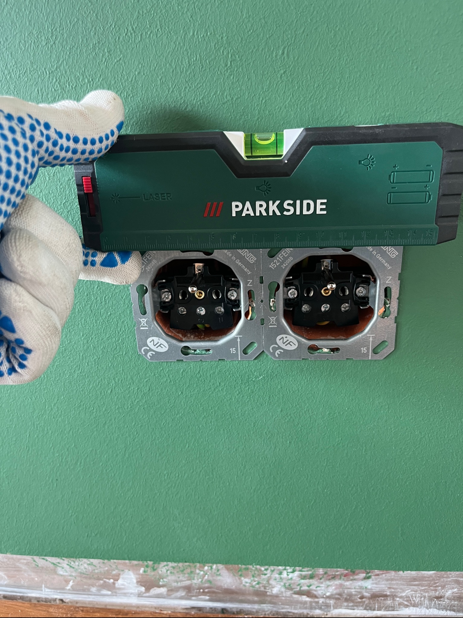 Elektryk w rękawicy w kropki poziomuje laserowym poziomem Parkside podwójne gniazdko elektryczne na zielonej ścianie.