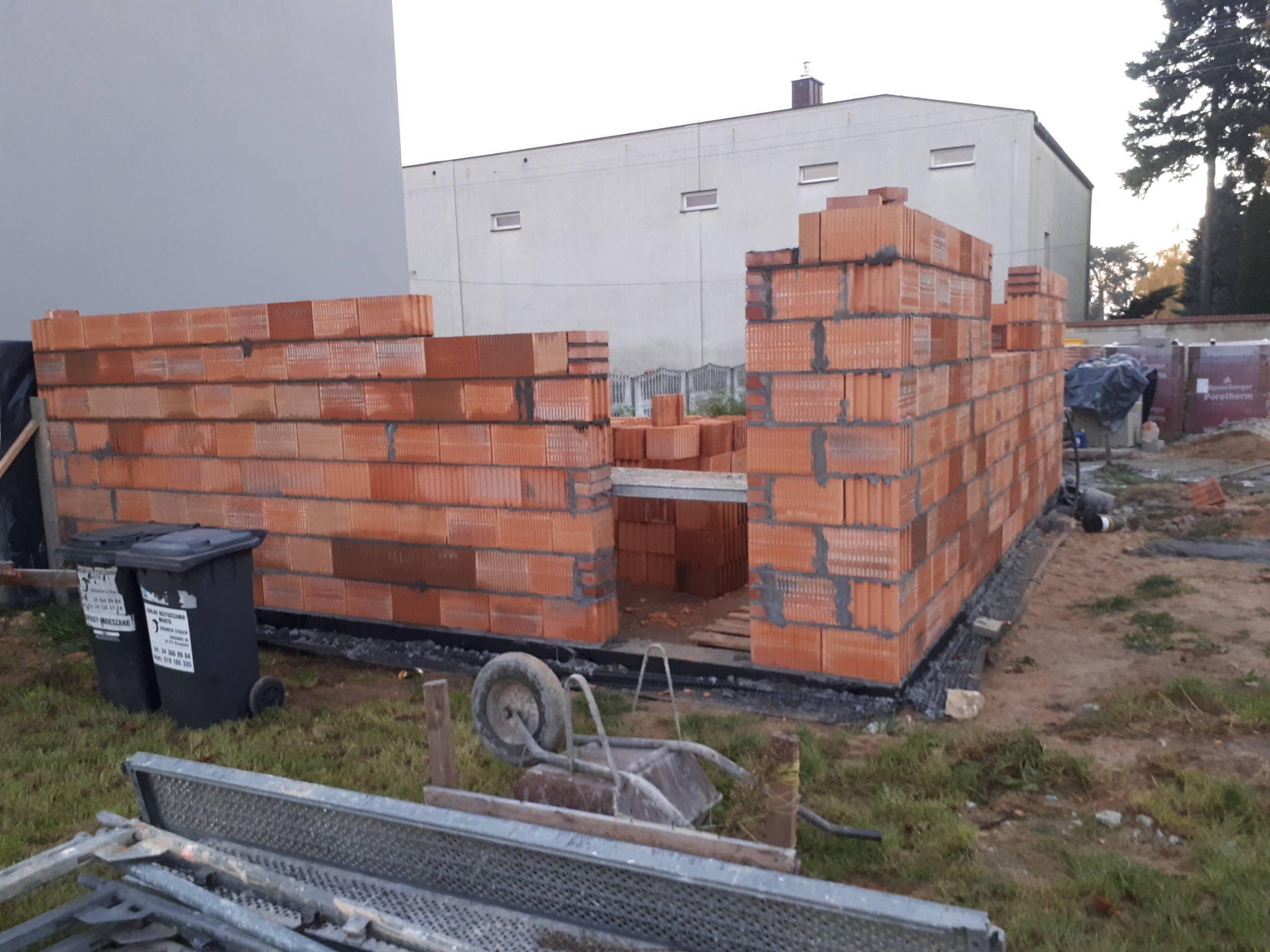 Wznoszenie ścian garażu dwustanowiskowego z czerwonej cegły ceramicznej Porotherm, widoczna hydroizolacja fundamentów, w tle budynek gospodarczy i fragment rusztowania.