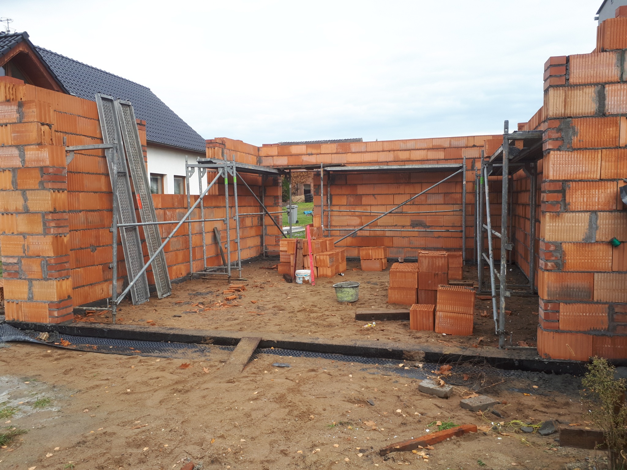 Wznoszenie ścian garażu z bloczków ceramicznych, widoczne rusztowania i materiały budowlane, w tle dom jednorodzinny.