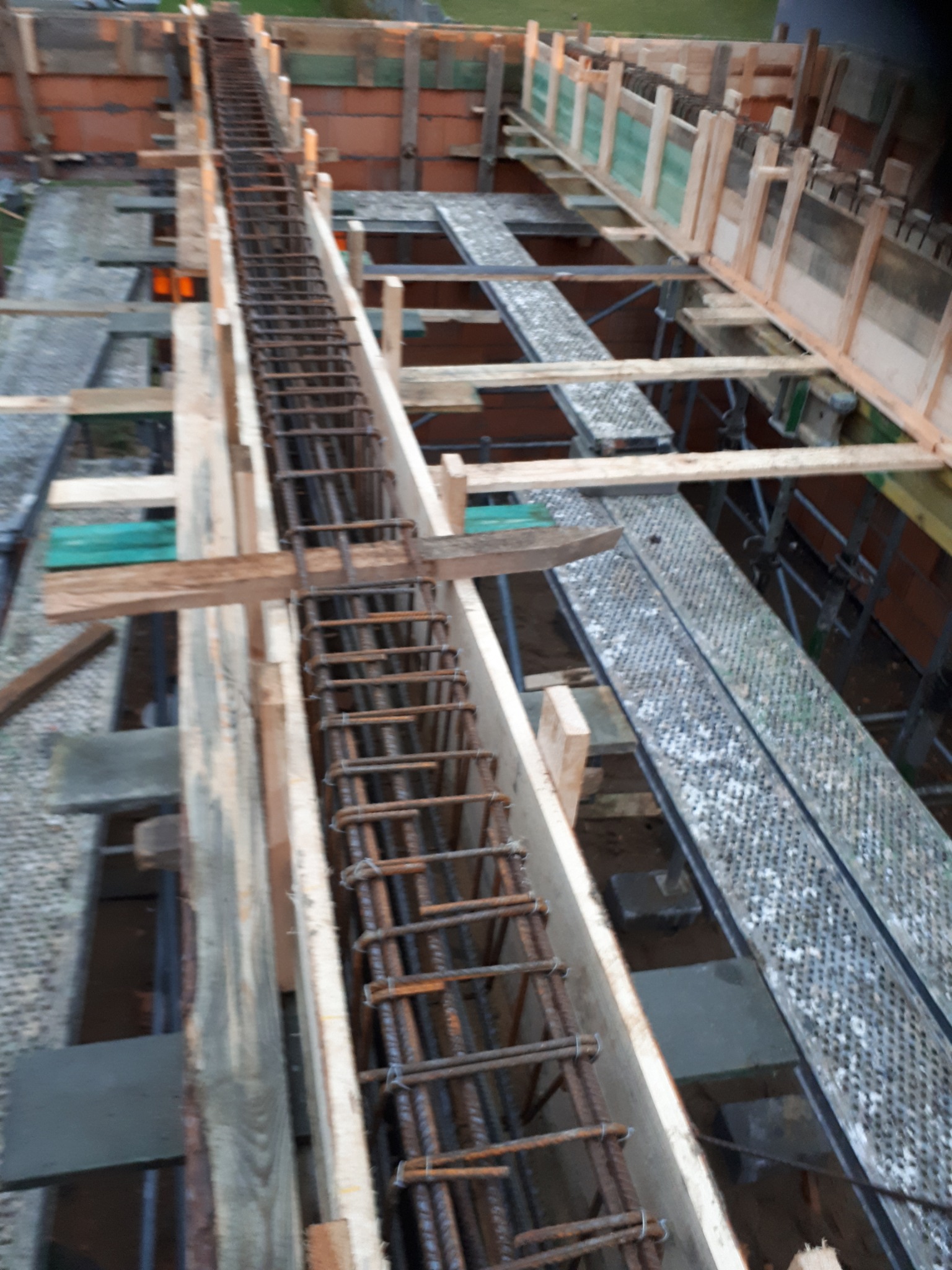 Zbrojenie w szalunku fundamentowym, widoczne pręty stalowe połączone drutem, drewniane deski tworzące formę, w tle rusztowanie i ceglany mur.