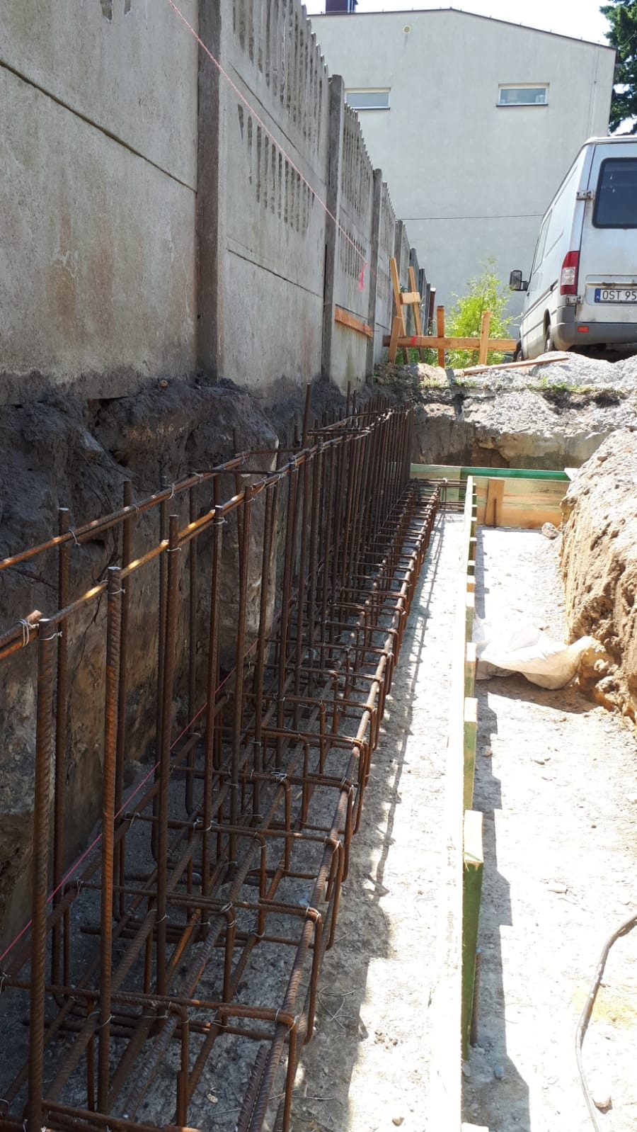 Zbrojenie fundamentów garażu dwustanowiskowego w wykopie, widoczna szalunek, w tle betonowy płot i biały bus
