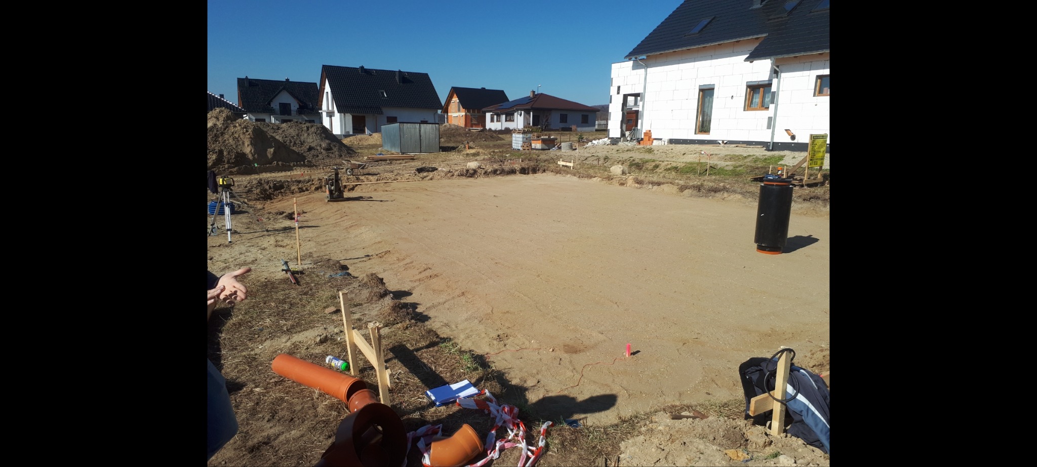 Przygotowany teren pod płytę fundamentową domu jednorodzinnego w Leśnicy, widoczne rury kanalizacyjne, tyczki geodezyjne oraz budynki w tle.