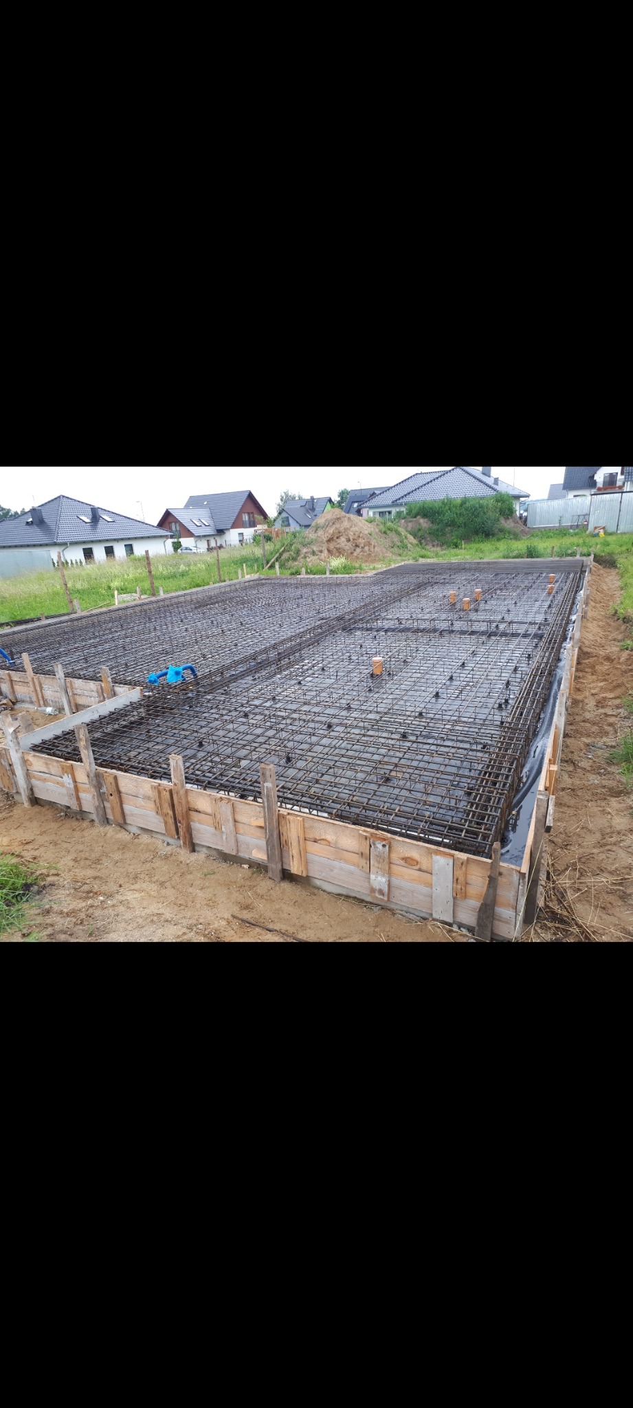 Przygotowana płyta fundamentowa zbrojona siatką stalową, otoczona drewnianym szalunkiem, z widocznymi pionowymi rurami kanalizacyjnymi i niebieskimi rurami instalacyjnymi, na tle domów...