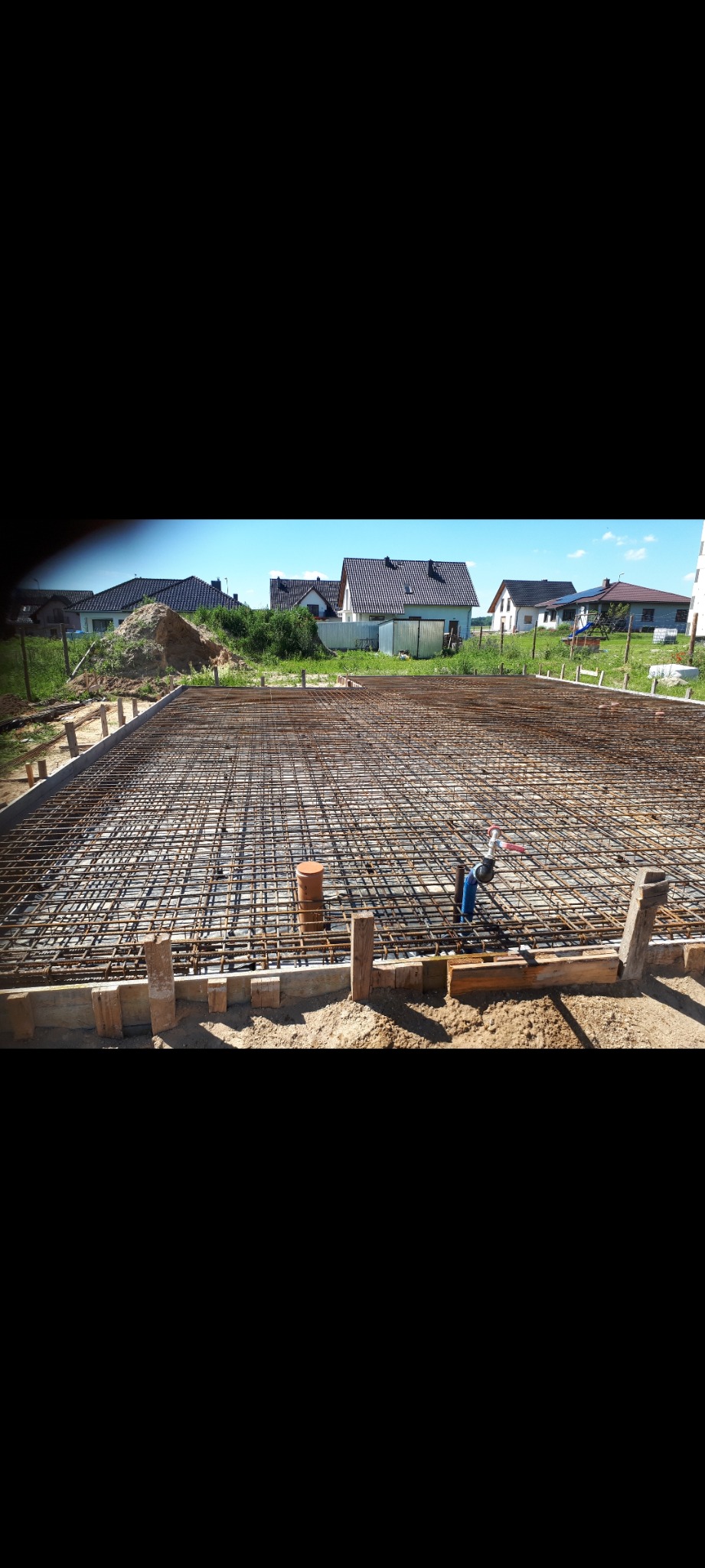 Zbrojenie płyty fundamentowej domu jednorodzinnego, widoczne szalunki drewniane, rury kanalizacyjne wystające ponad siatkę zbrojeniową oraz budynki mieszkalne w tle.