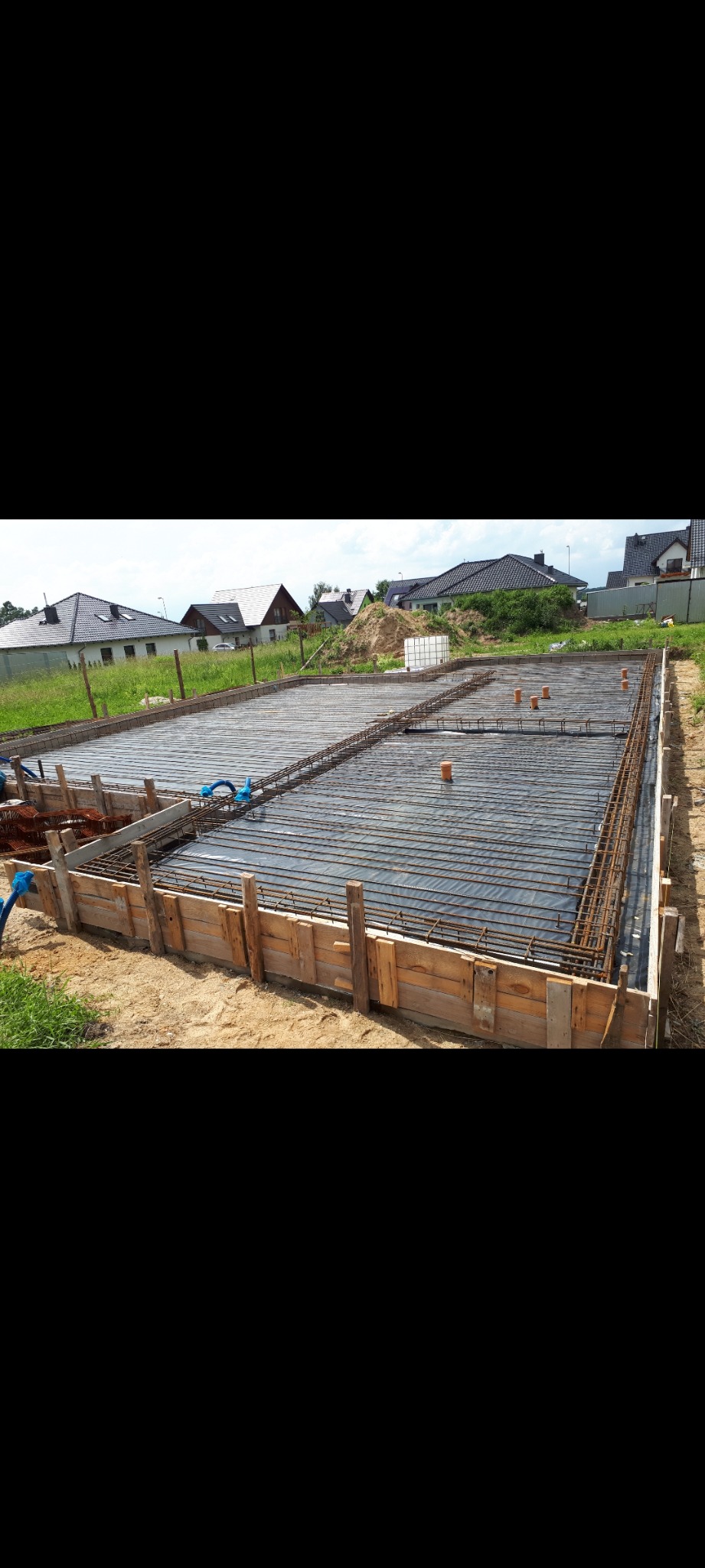 Przygotowana płyta fundamentowa z drewnianym szalunkiem, zbrojeniem i folią izolacyjną, widoczne pionowe rury kanalizacyjne, w tle zabudowa jednorodzinna.