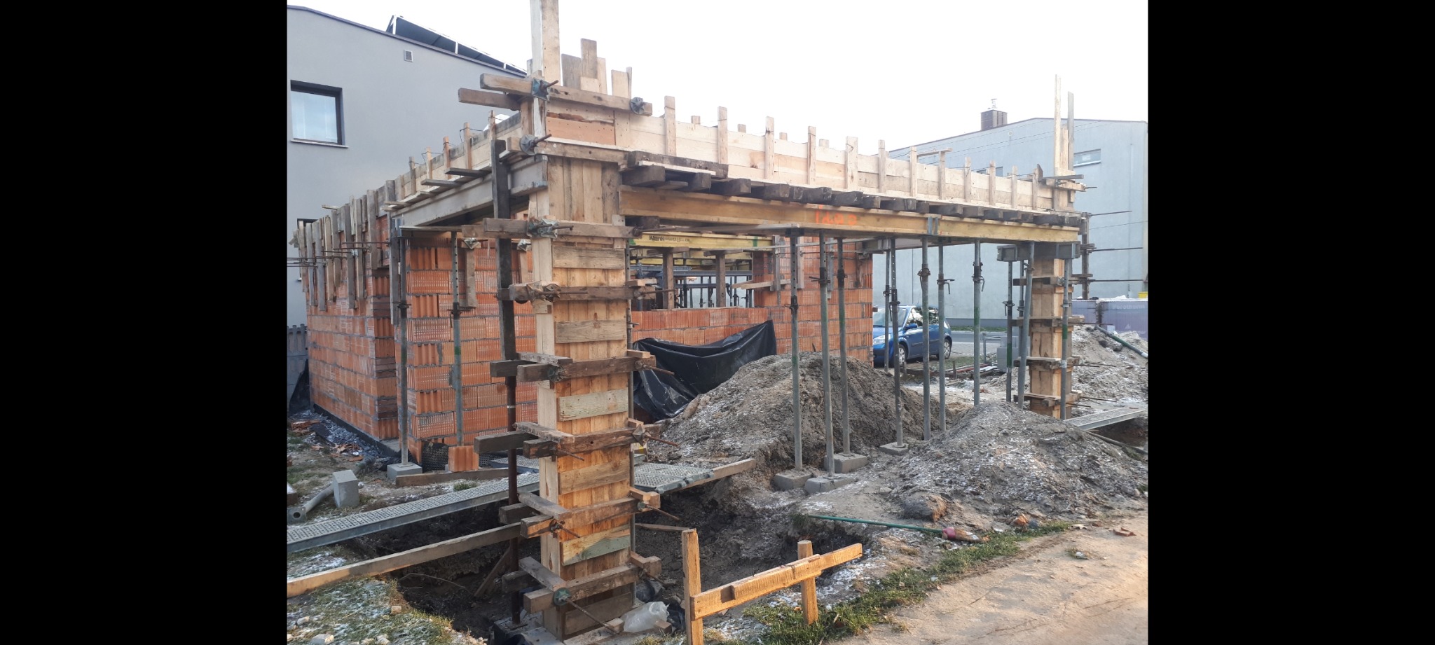 Budowa dwustanowiskowego garażu z cegły z drewnianym szalunkiem i podporami, widoczny wykop pod fundamenty, w tle zabudowa mieszkalna.