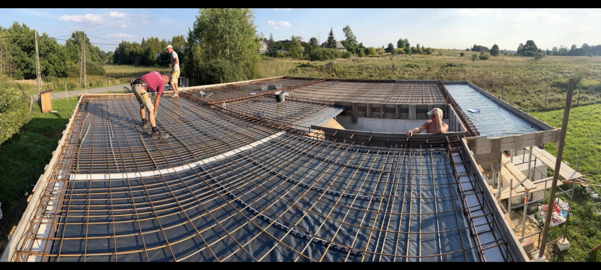 Zbrojenie stropu domu jednorodzinnego w trakcie realizacji: widoczne stalowe pręty, folia izolacyjna, szalunki i pracownicy budowlani na tle zielonego krajobrazu.