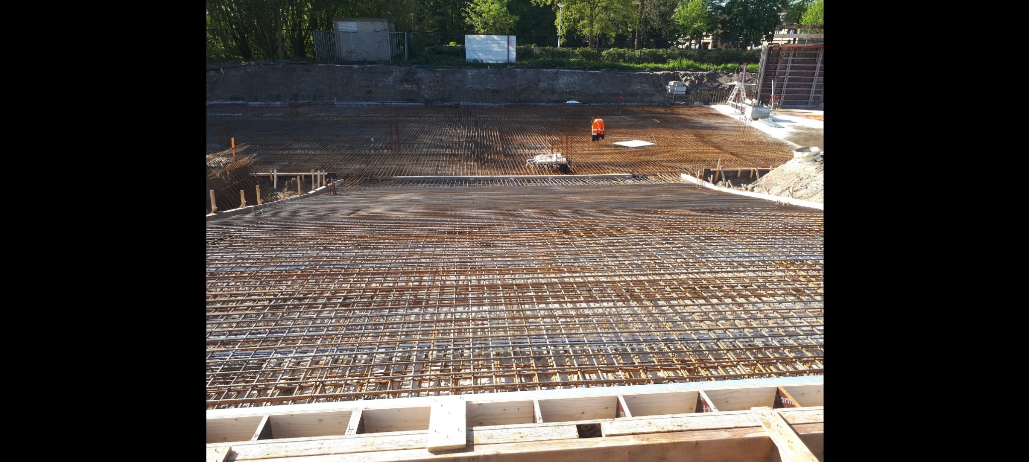 Zbrojenie płyty fundamentowej z widoczną siatką stalową i szalunkiem drewnianym, w tle pracownik w pomarańczowej kamizelce.