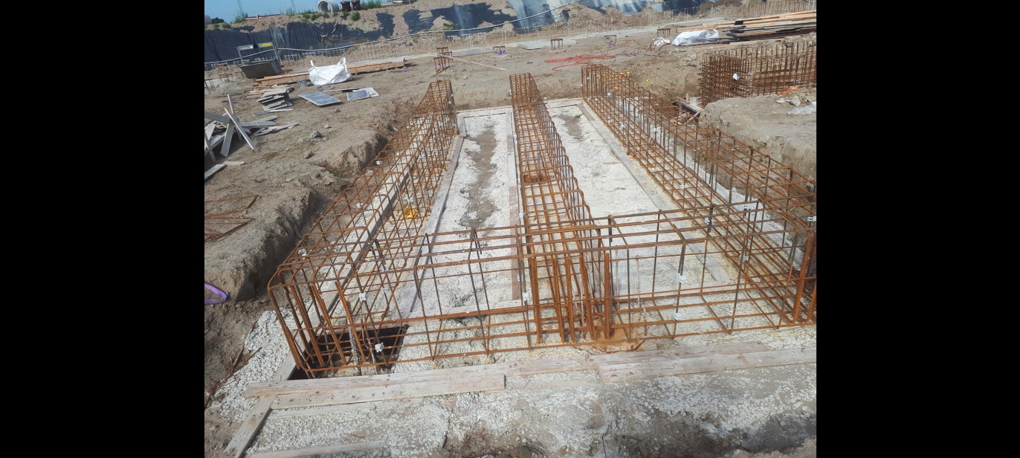 Zbrojenie fundamentów w wykopie, widoczne stalowe pręty tworzące szkielet konstrukcji, częściowo zalane białym betonem, przygotowane do dalszych prac budowlanych.