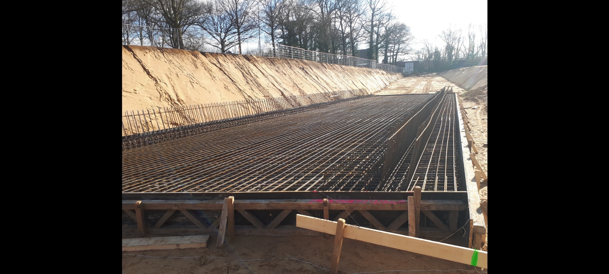 Zbrojenie fundamentów w wykopie, widok z góry na siatkę z prętów stalowych i drewniane szalunki, na tle skarpa ziemna i drzewa.