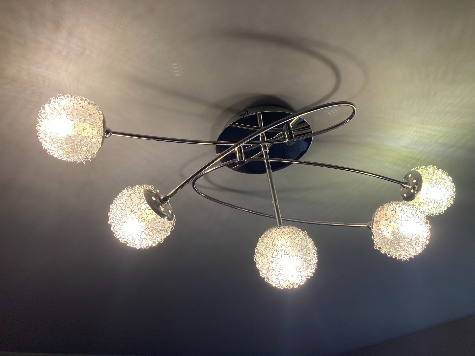 Nowoczesna lampa sufitowa z pięcioma kulistymi kloszami o ażurowej strukturze, zawieszona na chromowanych ramionach, rzucająca cienie na sufit.