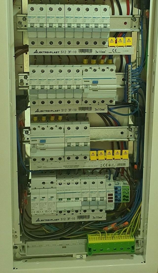 Rozdzielnica elektryczna w białej obudowie, widoczne zabezpieczenia, przewody i listwy zaciskowe. Instalacja połączona, gotowa do użytku.