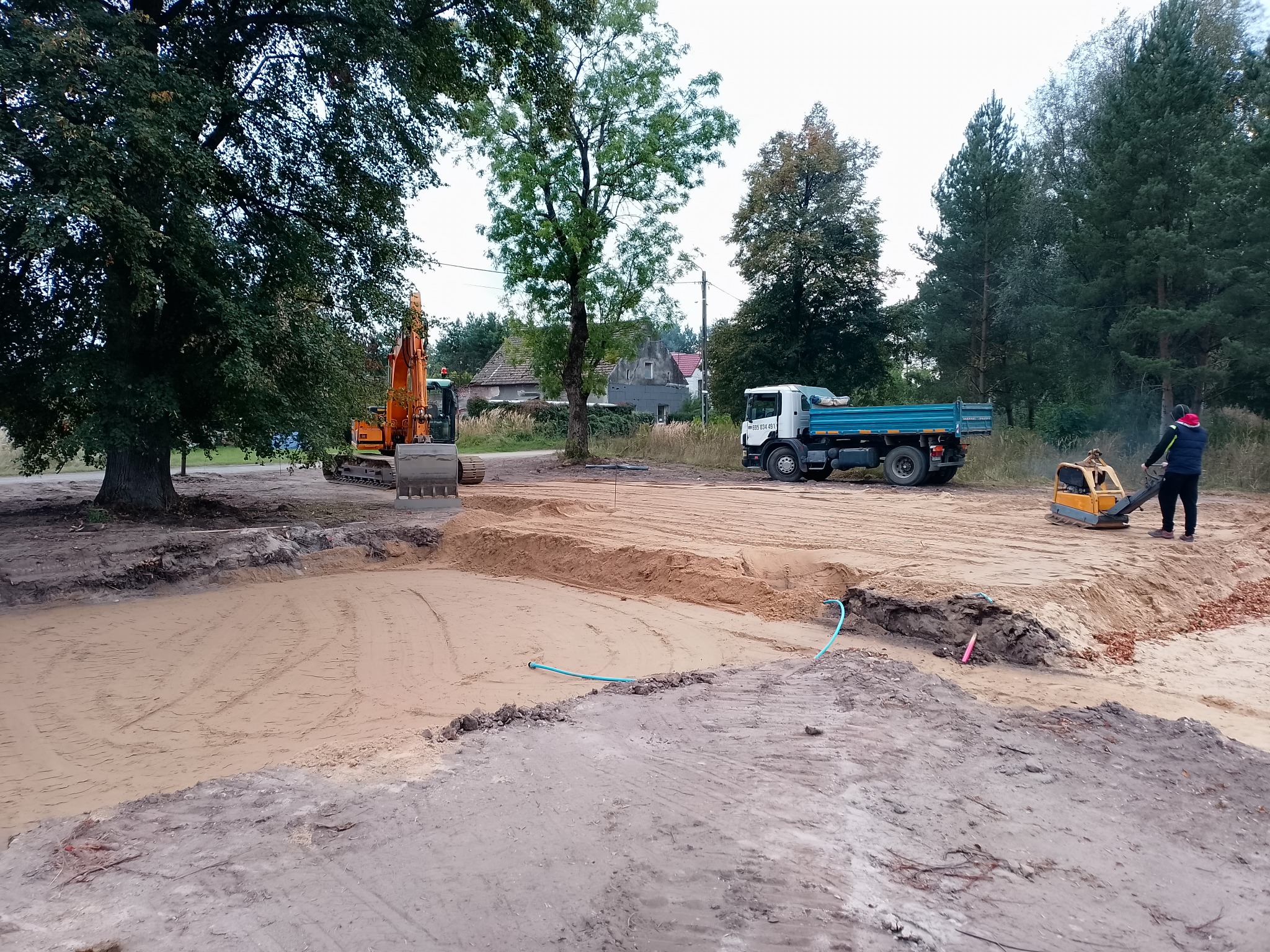 Prace ziemne: koparka, wywrotka i zagęszczarka na placu budowy. Widoczny wykop z piaskiem, przygotowany teren pod budowę. Niebieski wąż na ziemi.